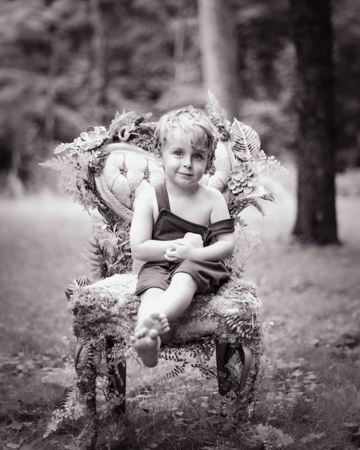 Black and white photo of a child sitting in a rustic floral chair outdoors.