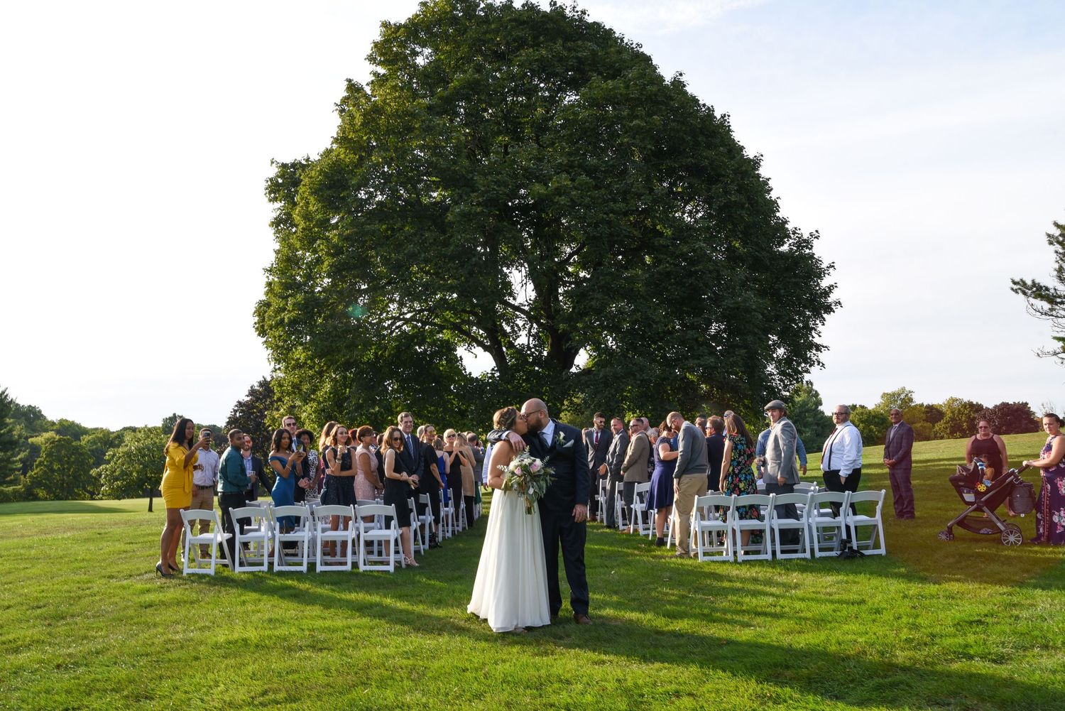 Panoramic view of outdoor wedding ceremony setup with white chairs on grassy hillside.