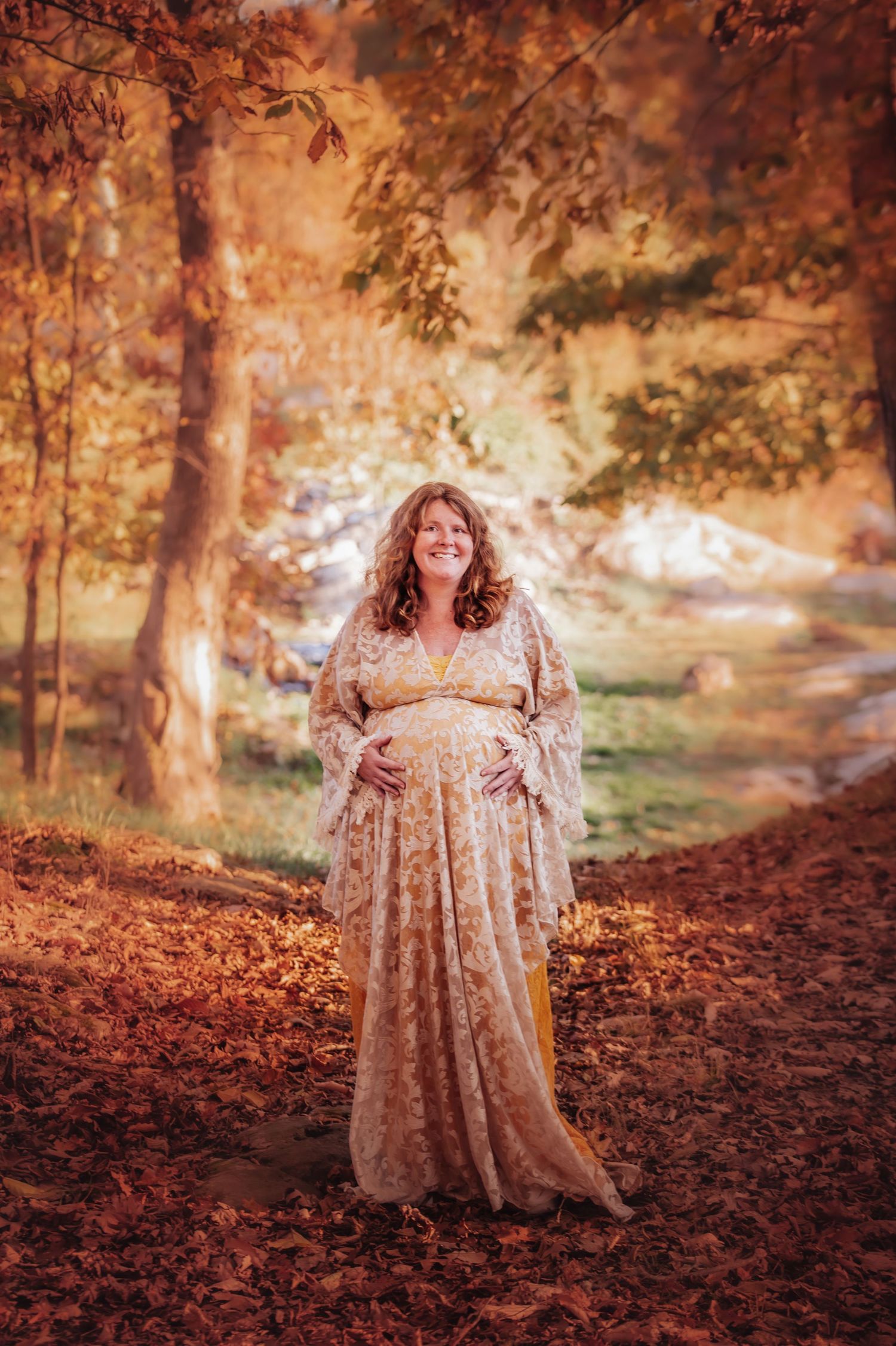 A dreamy autumn maternity photo featuring a flowing lace dress against a backdrop of golden fall foliage and sunlit trees.
