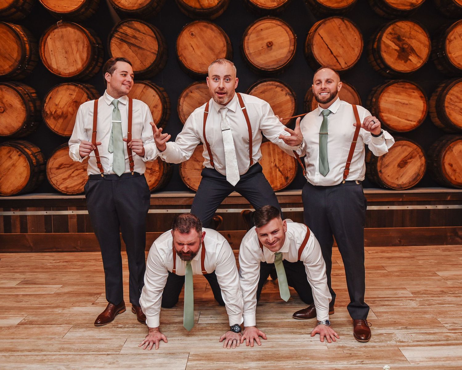 Groomsmen in suspenders and bowties posing playfully on wooden floor.