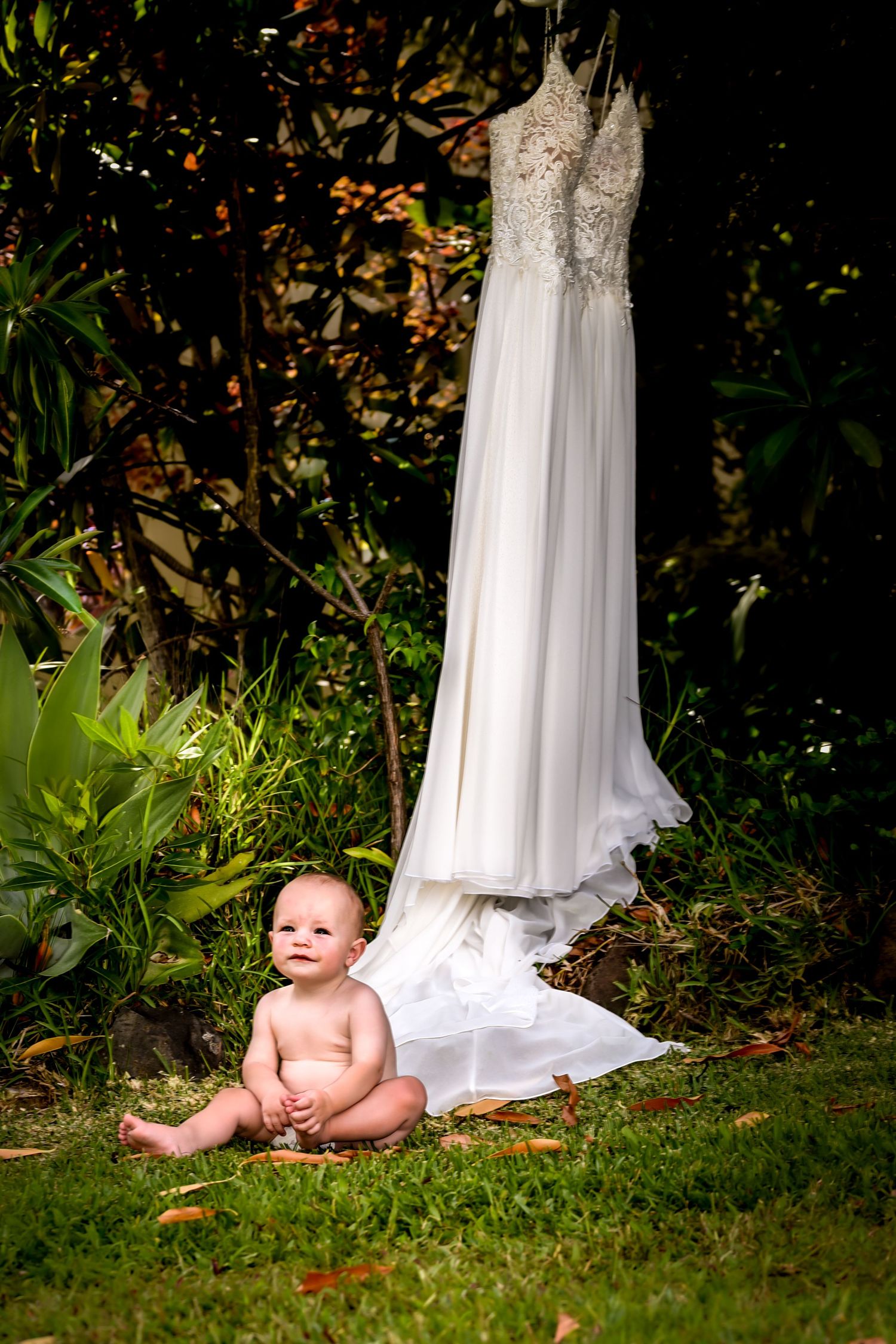 White wedding dress hangs elegantly against autumn foliage while a baby plays on grass nearby.