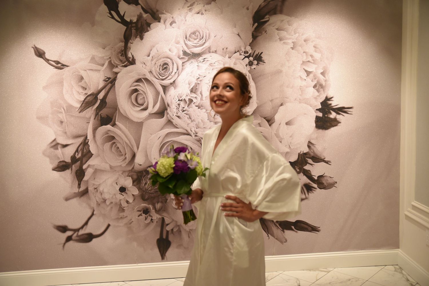 A white wedding dress hangs against an elegant floral wallpaper background.