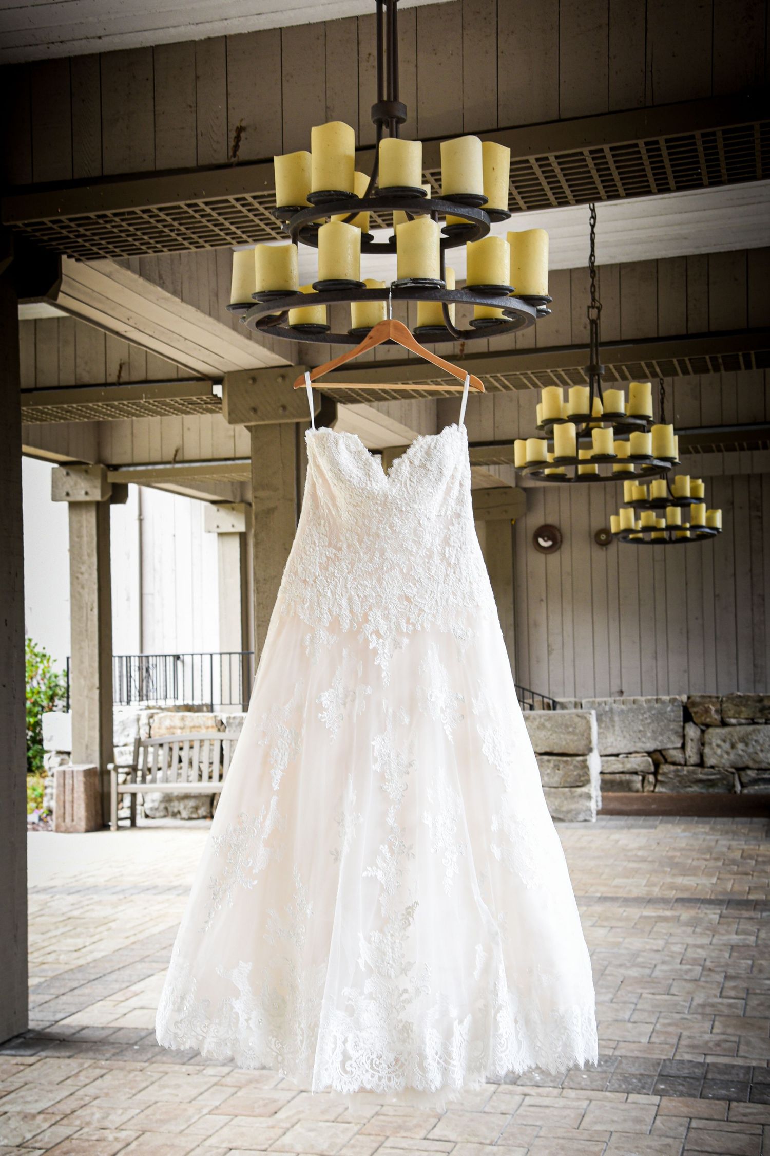 A white lace wedding dress hangs elegantly from a wooden hanger beneath rustic chandeliers in a covered porch setting.