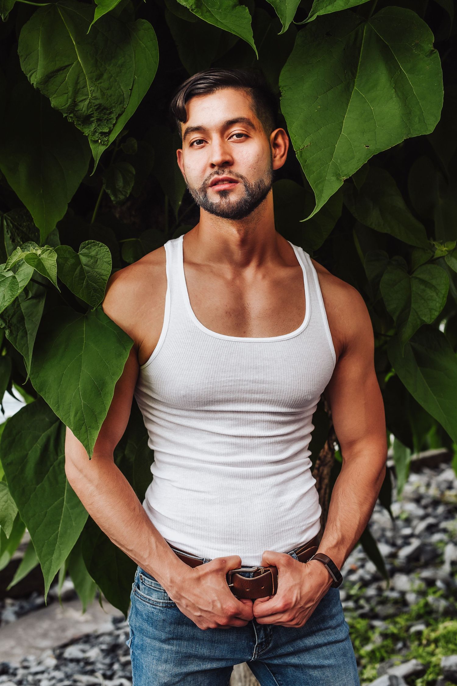Athletic figure in white tank top and jeans poses against lush green foliage backdrop in outdoor photoshoot.