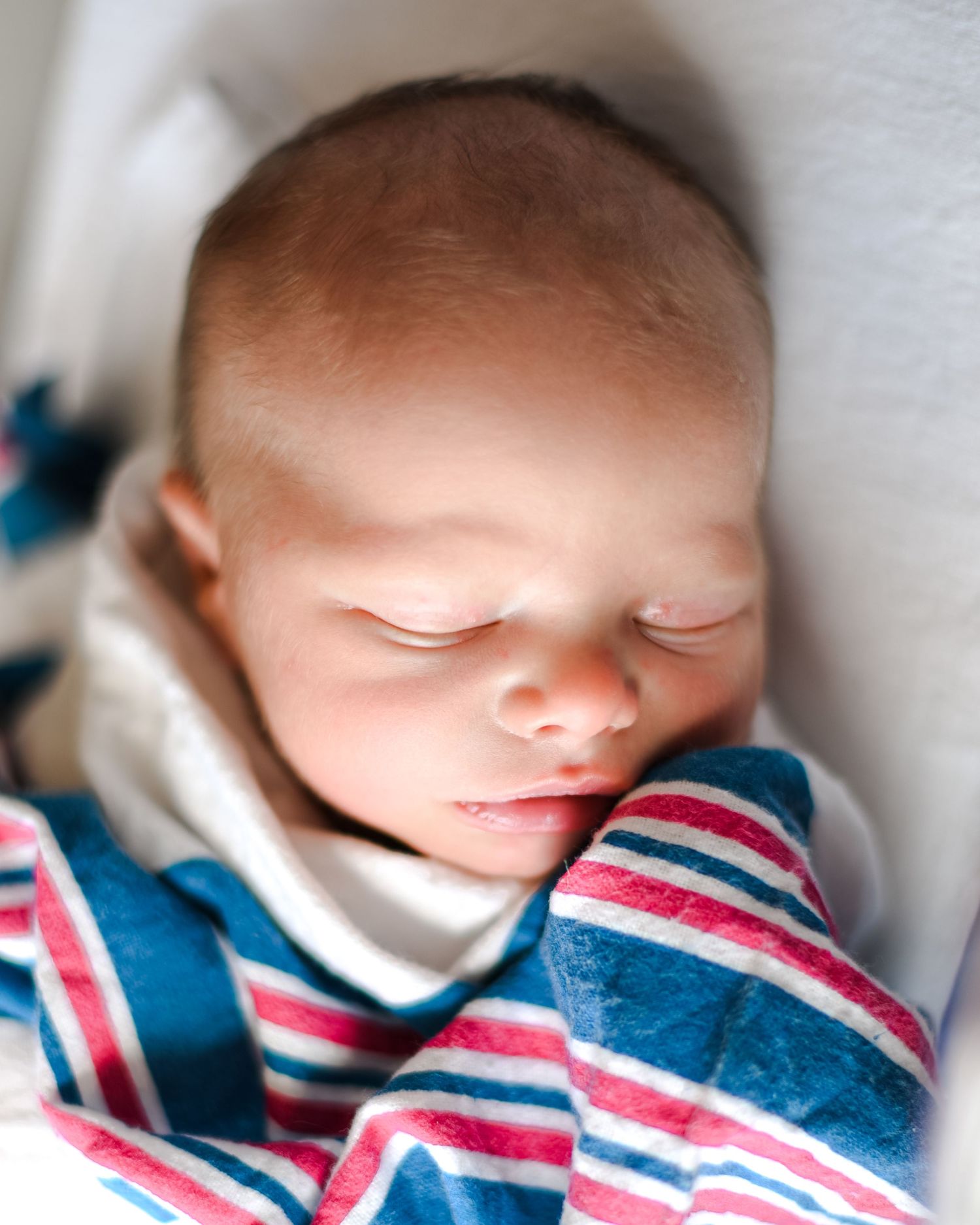 Sleeping baby wearing a striped red and blue shirt rests peacefully on white bedding.
