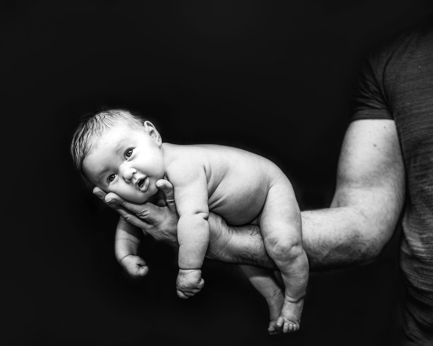 Black and white artistic portrait of a baby being held horizontally against dark background.