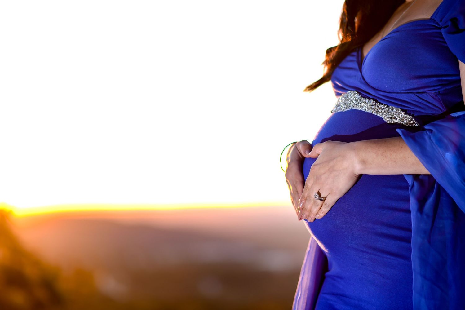 Silhouette of a pregnant woman in a blue dress cradling her belly against a warm sunset background.