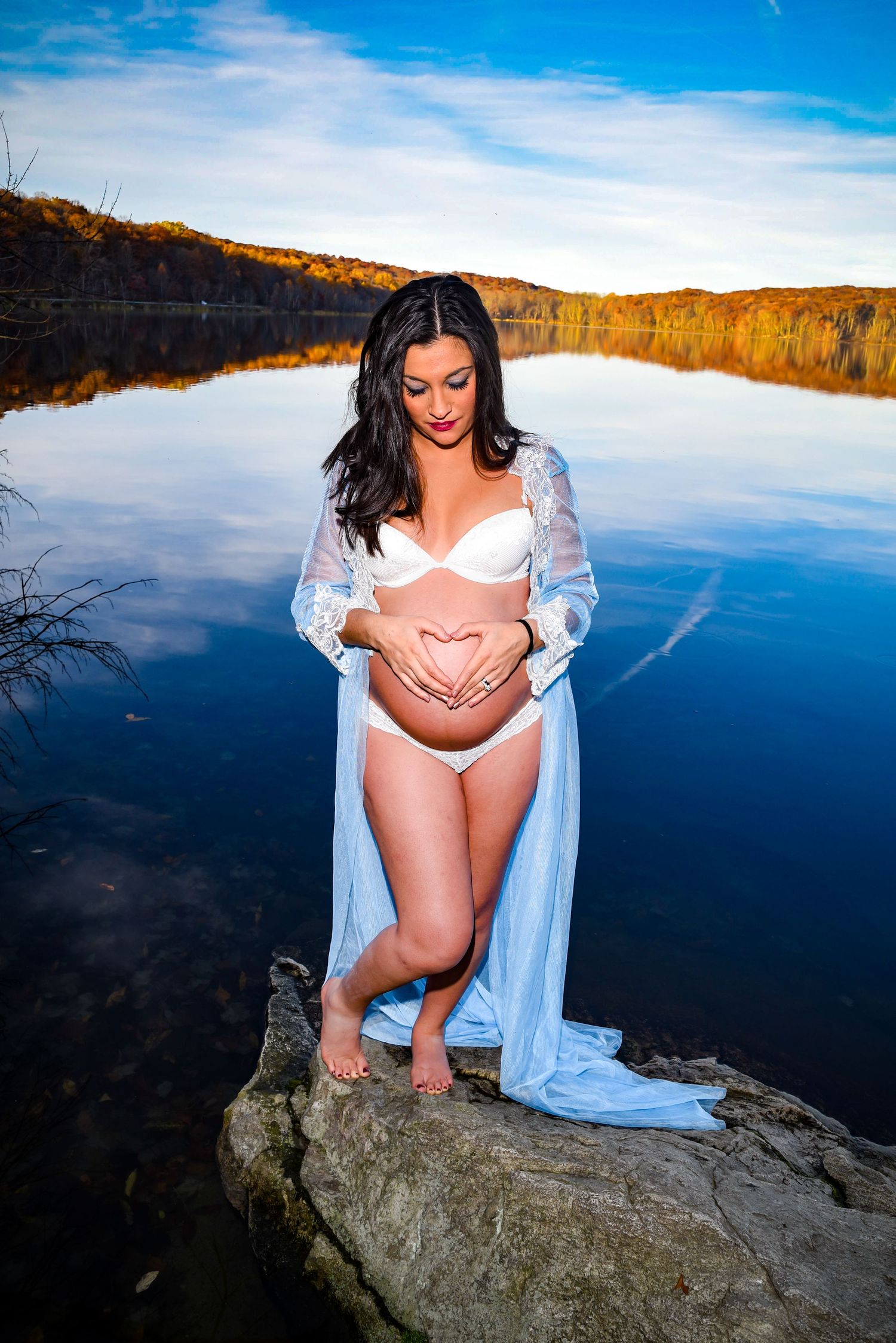 Artistic maternity photoshoot at a scenic lake overlook with dramatic blue sky reflections and rocky outcrops.
