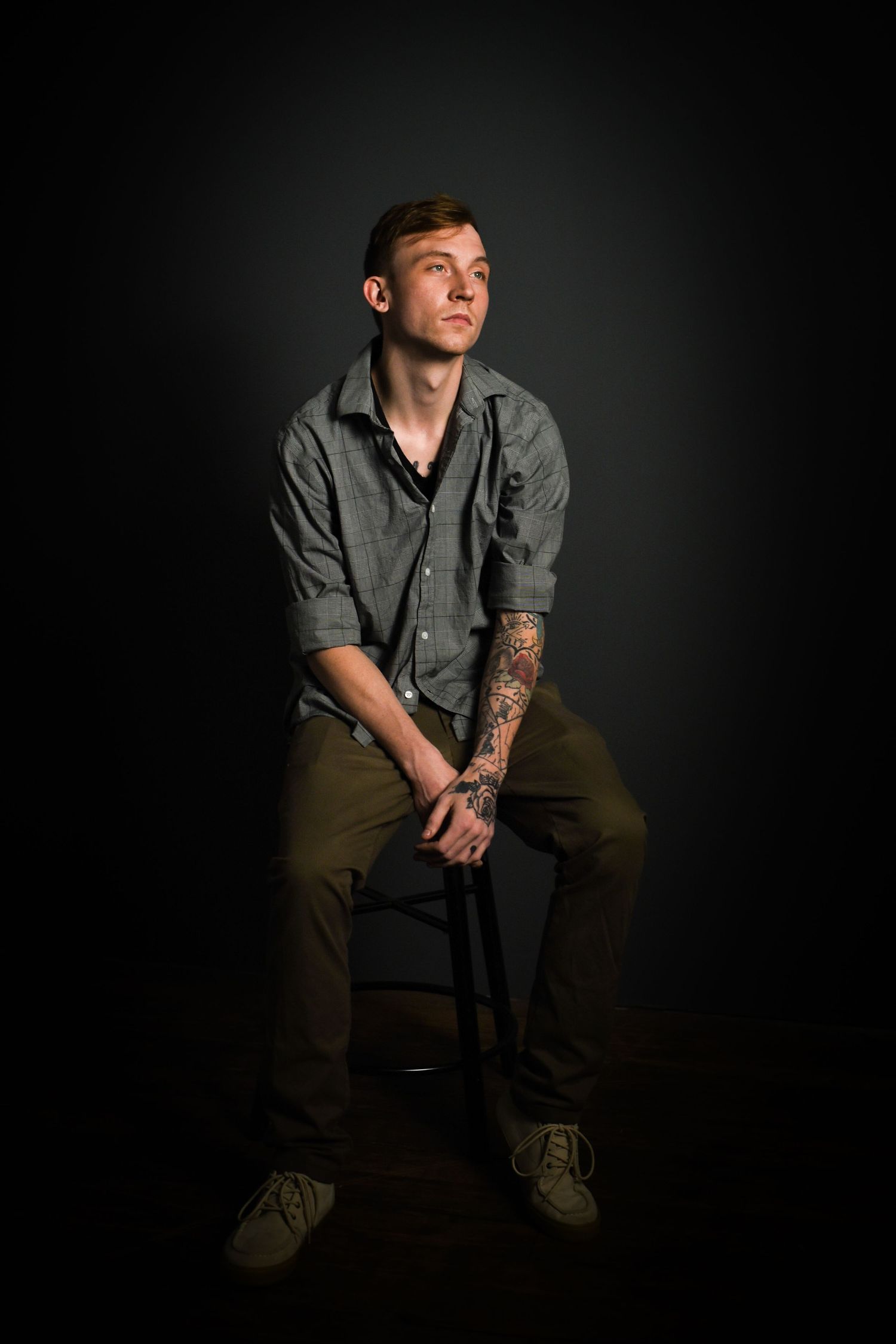 Moody studio portraits of person in grey shirt and dark pants seated against black backdrop.