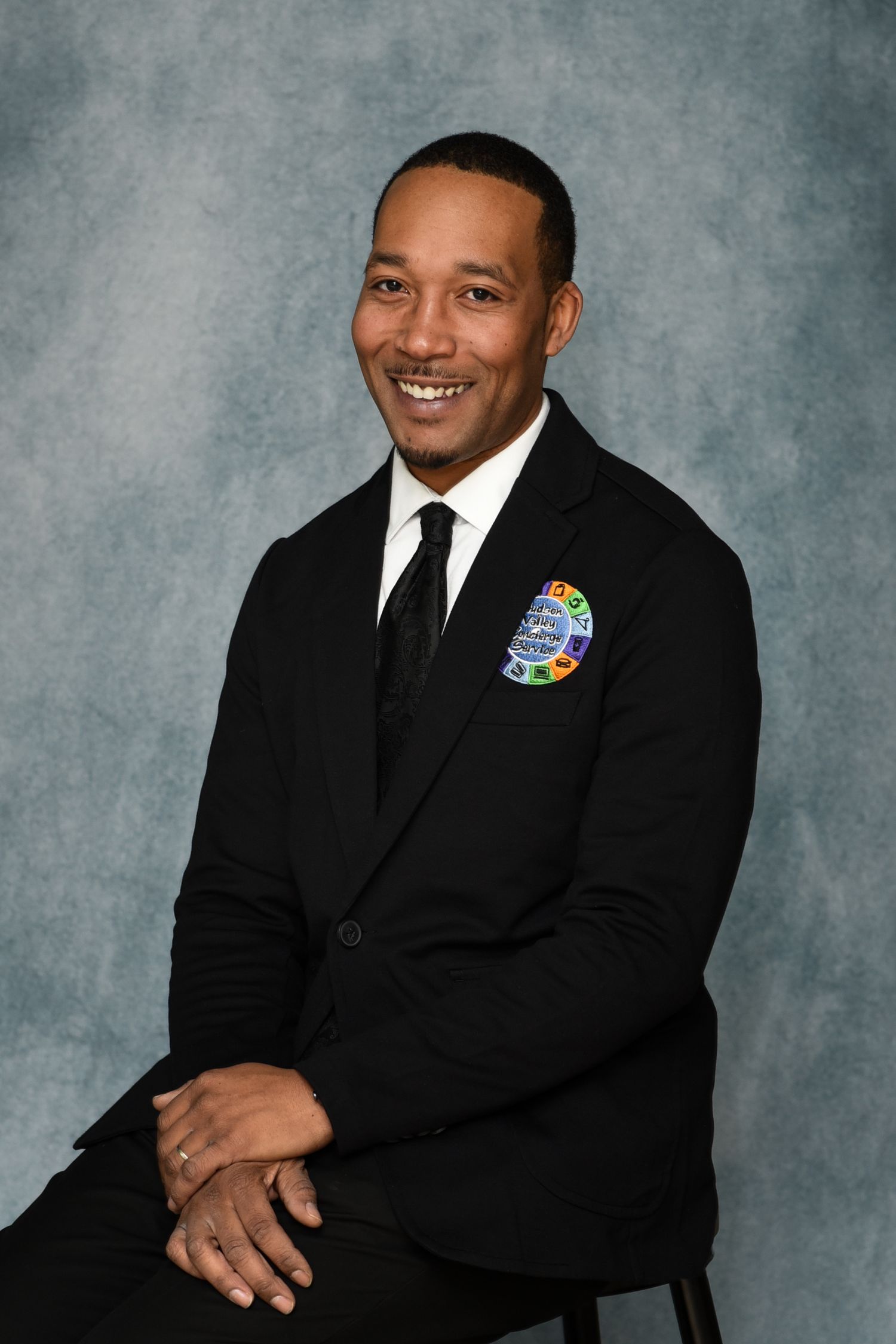 Professional portrait of a person in a black suit and tie with a colorful pin on their lapel against a gray background.