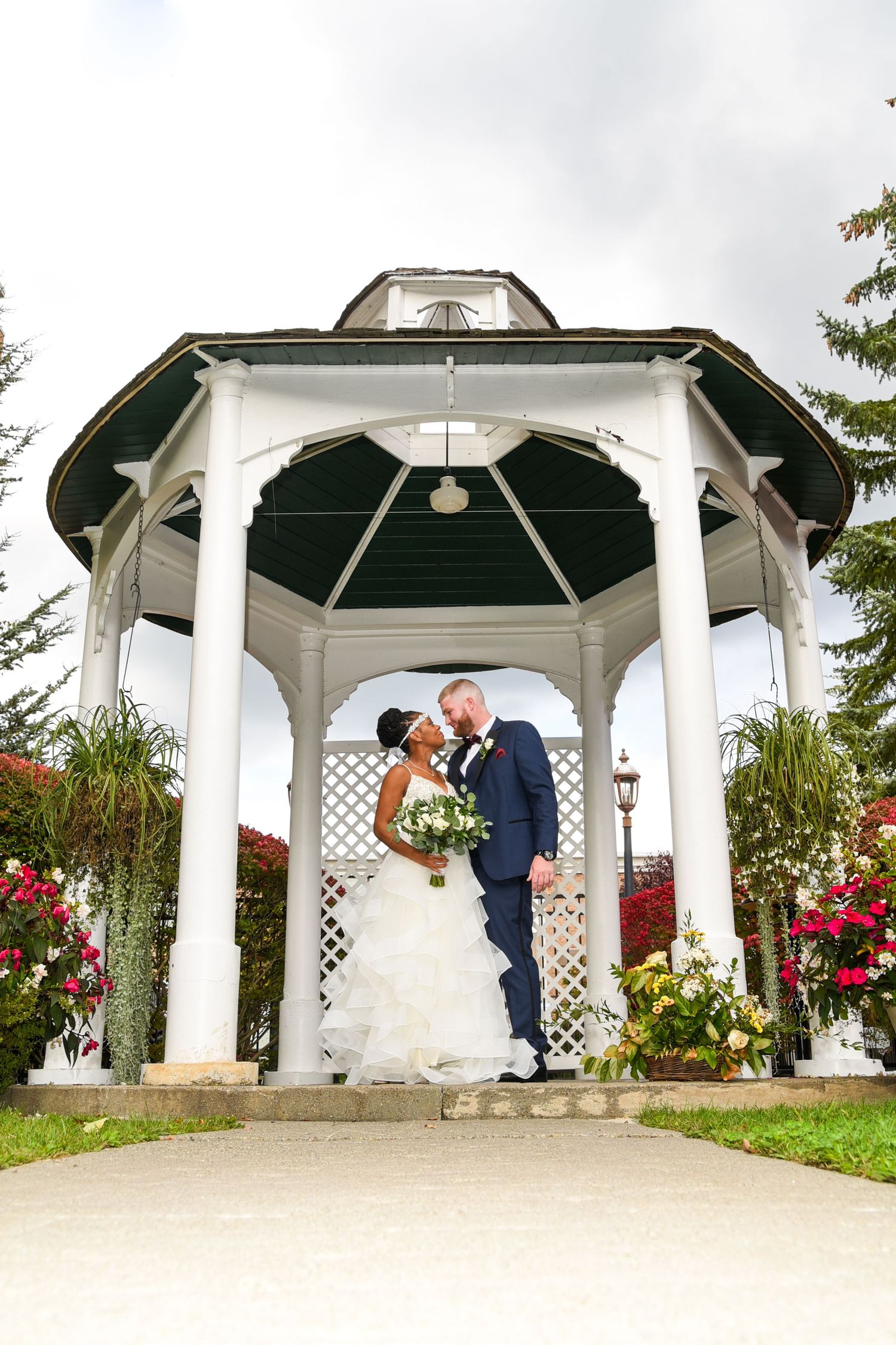 Newlyweds share romantic moment in white gazebo surrounded by red flowers.