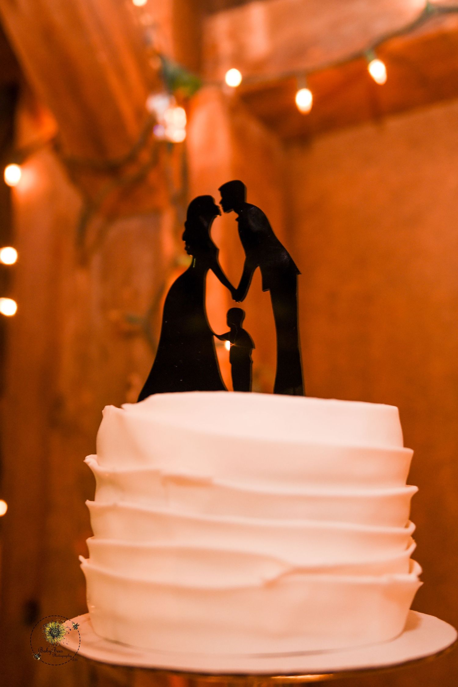 White wedding cake with silhouette topper illuminated by warm string lights in rustic venue setting.