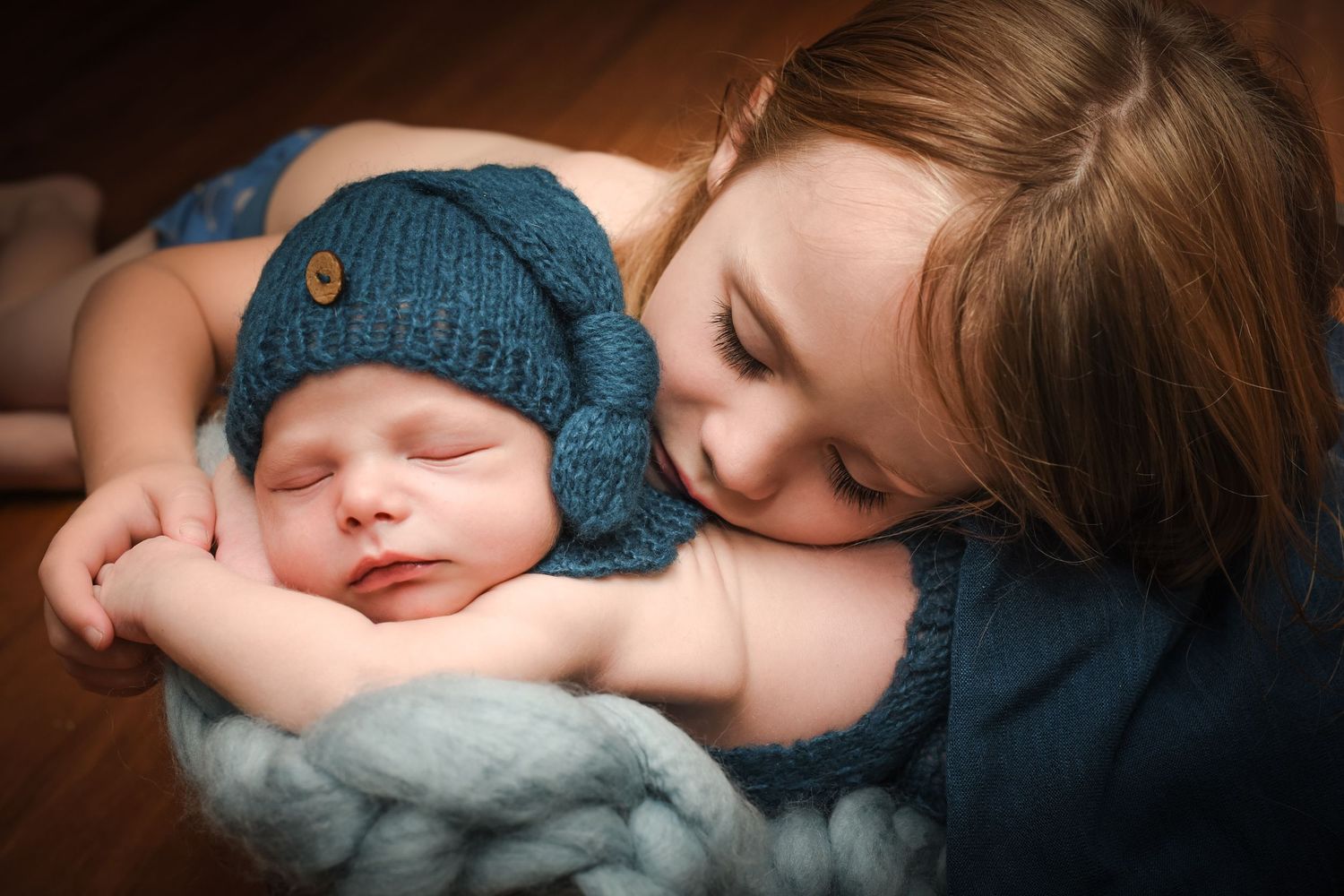 A sleeping newborn wearing a blue knit hat is gently embraced while resting on a soft blanket.