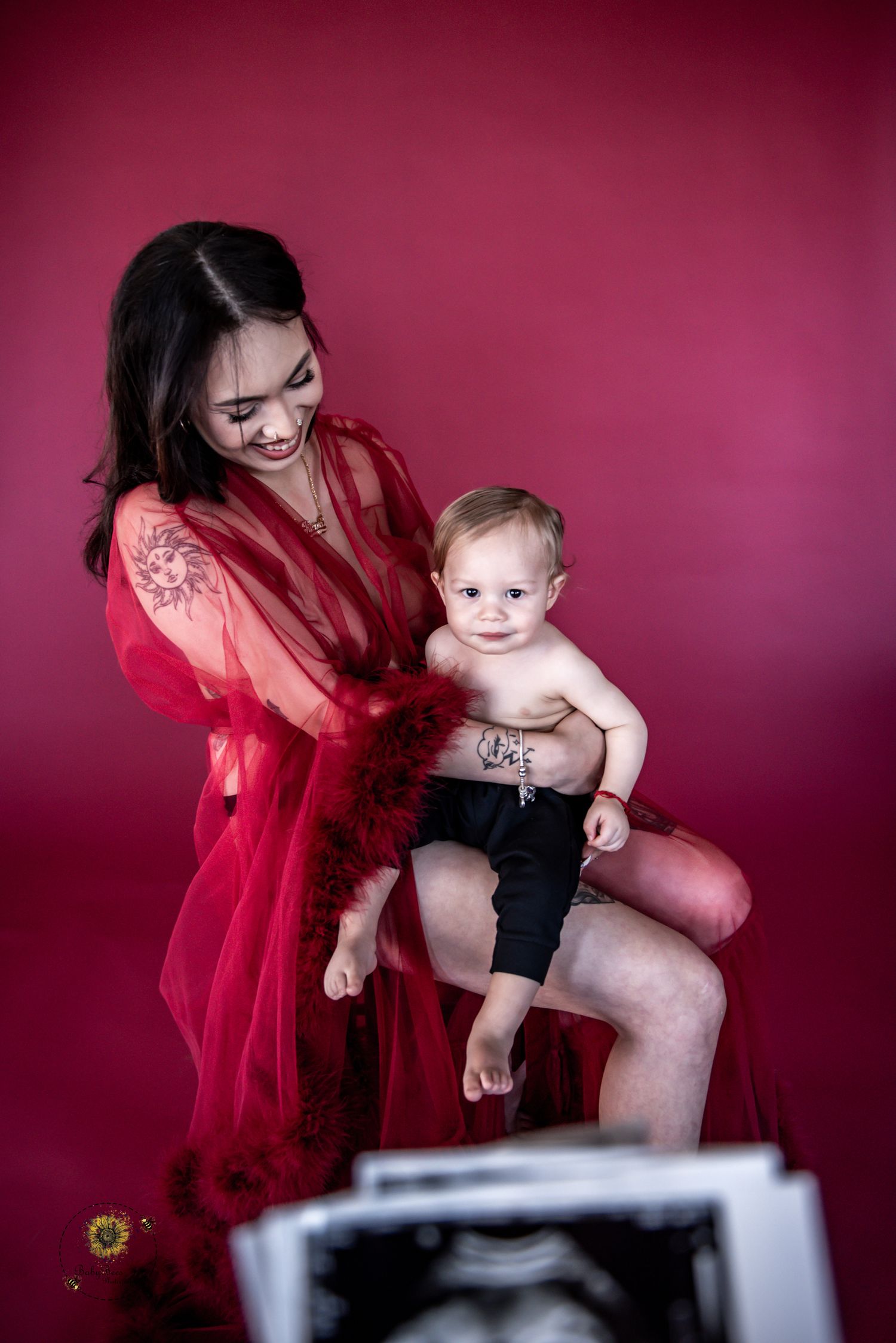 Intimate family portrait in red tones showing tender moment between parent and baby against burgundy backdrop.
