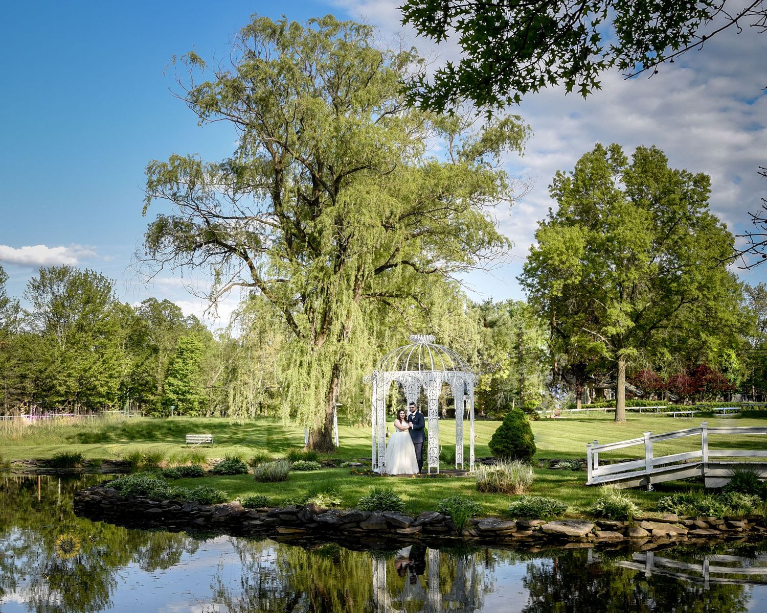 A wedding ceremony setup by a tranquil pond with a weeping willow tree and white gazebo.