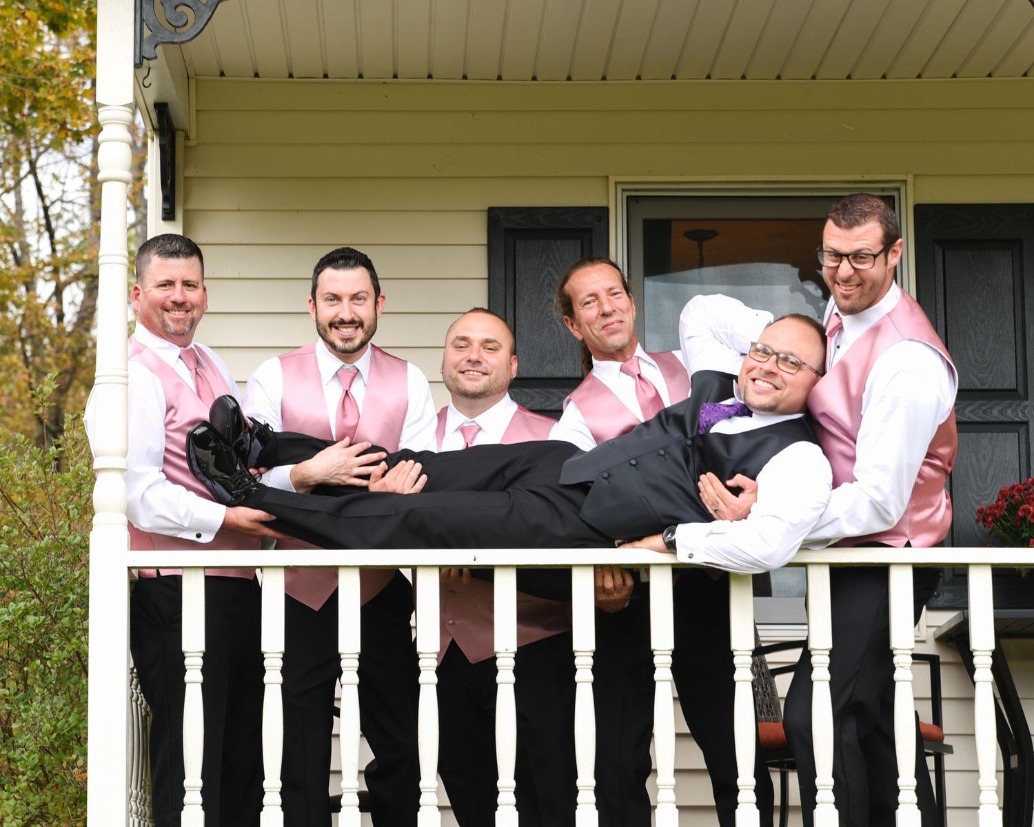 Wedding party poses together on white porch railing of country house.