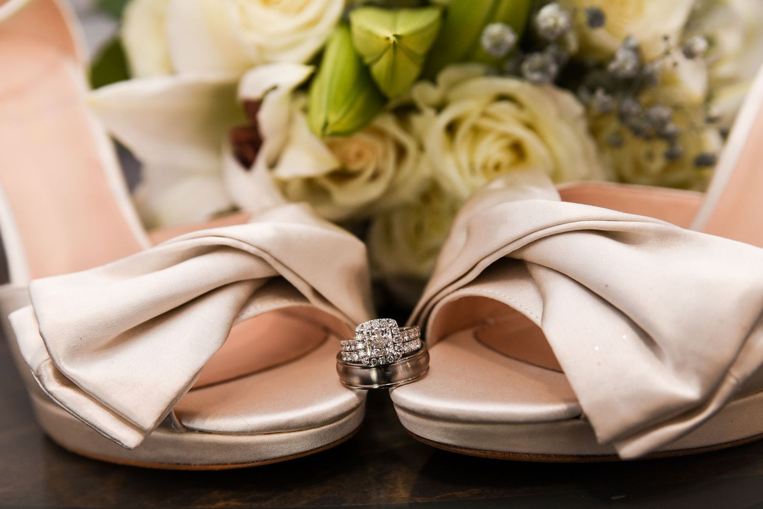 Elegant white satin bridal shoes with bow detail and diamond engagement ring displayed with roses in background.