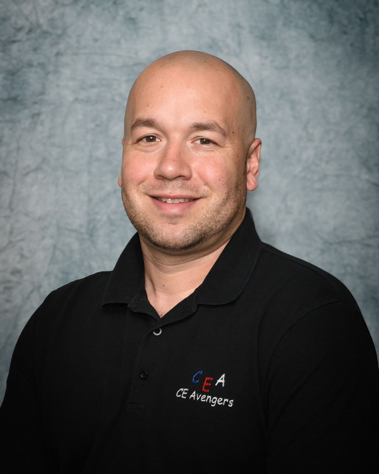 Professional headshot of a bald person in a black polo shirt with company logo against a gray background.
