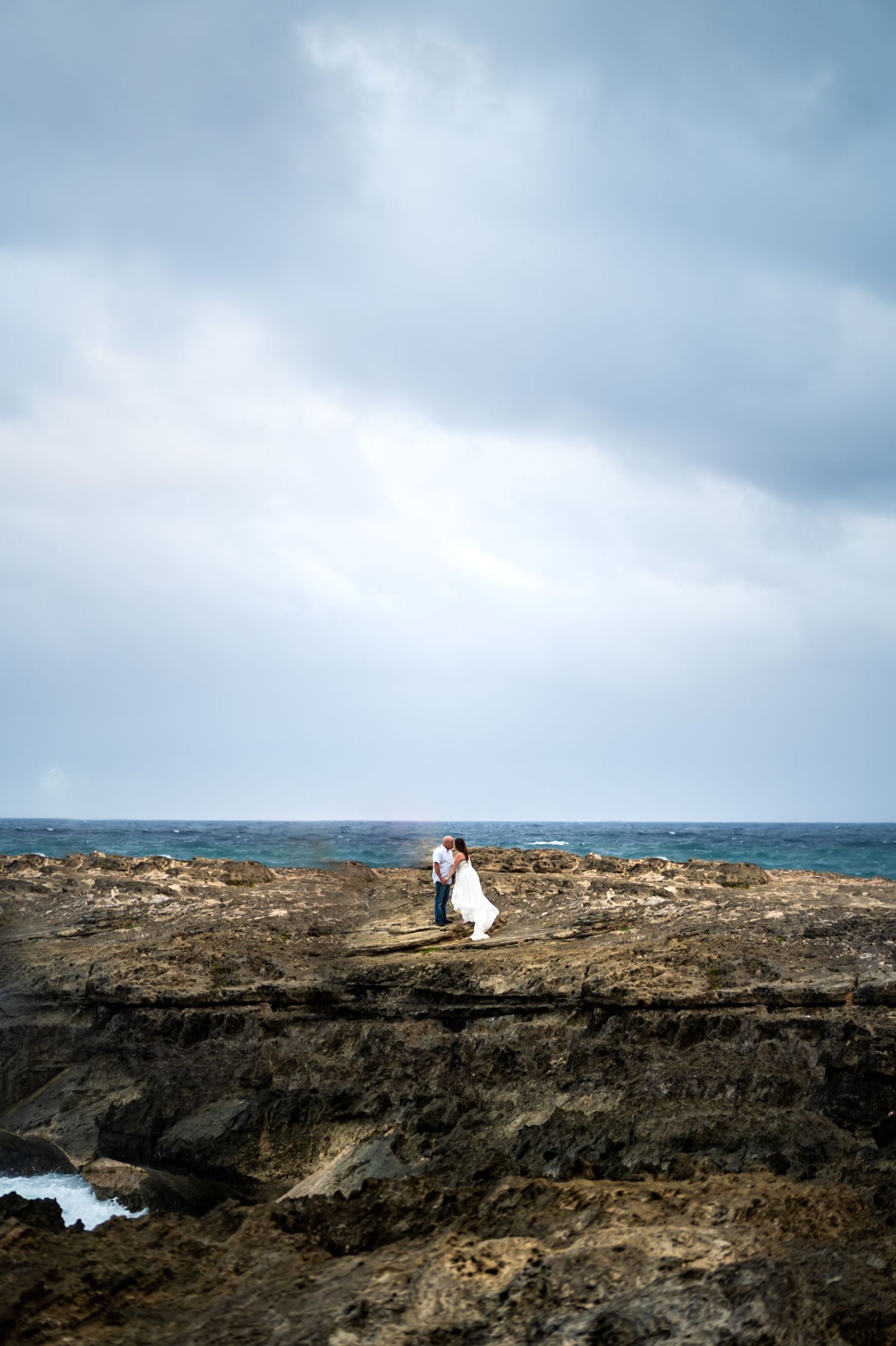 Dramatic rocky coastline with ocean waves crashing against cliffs under cloudy sky.