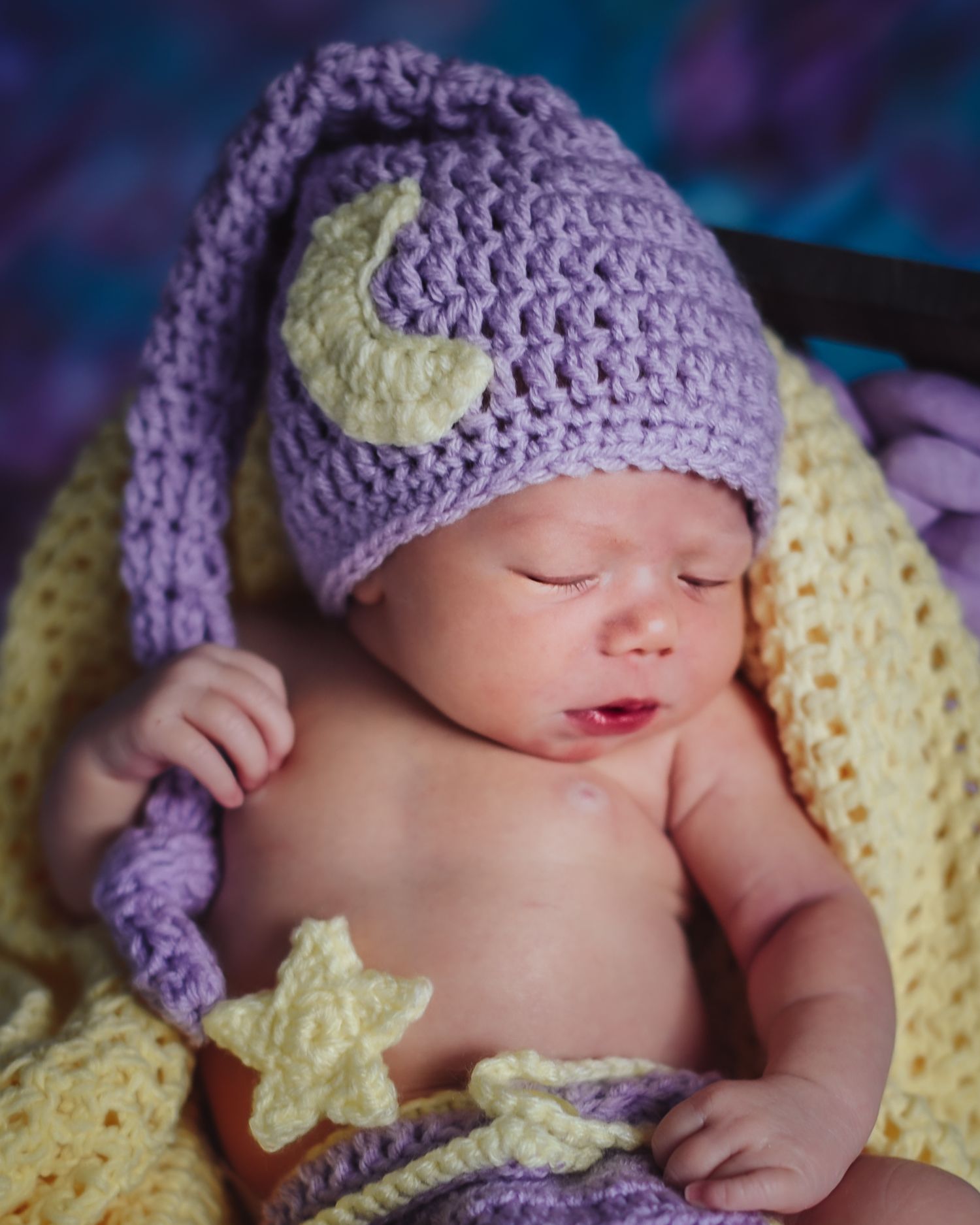Sleeping baby wearing purple crocheted nightcap with moon and star details lies on yellow blanket in dreamy newborn photo.