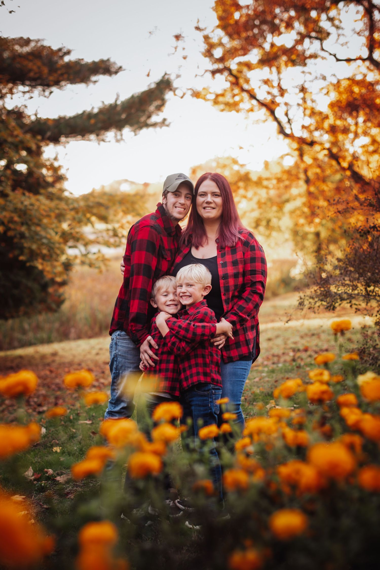 Family wearing matching red plaid flannel shirts pose together in autumn field with orange marigolds at sunset.
