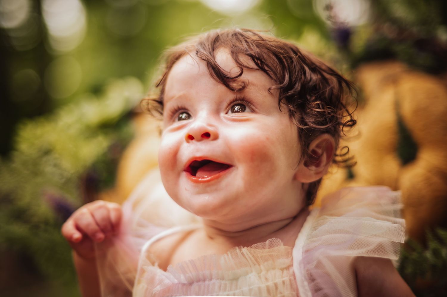 A joyful baby with curly hair smiles brightly in a white dress against a blurred natural background.