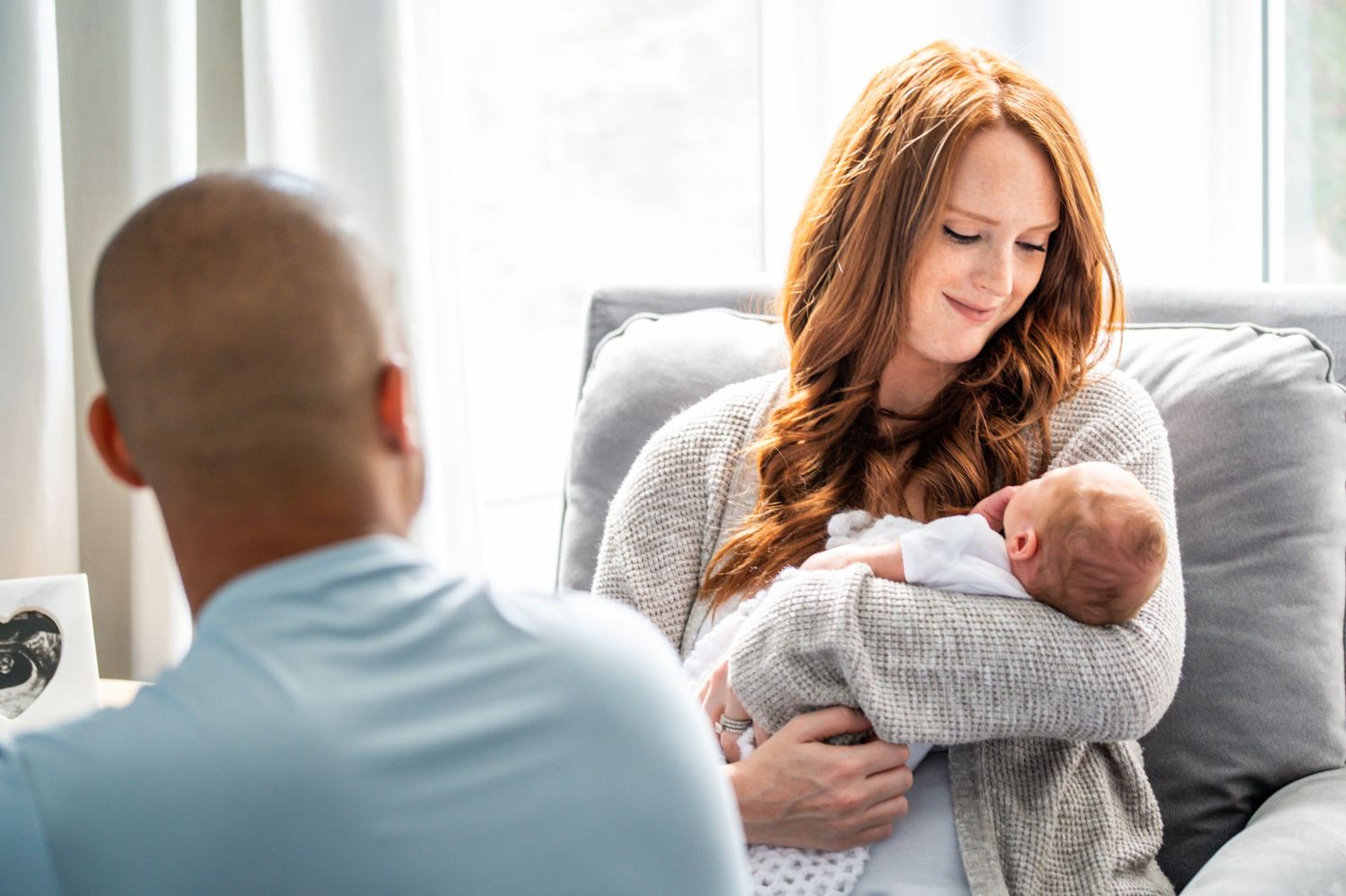 A peaceful moment at home as a parent holds a sleeping newborn baby while sitting on a cozy gray couch.