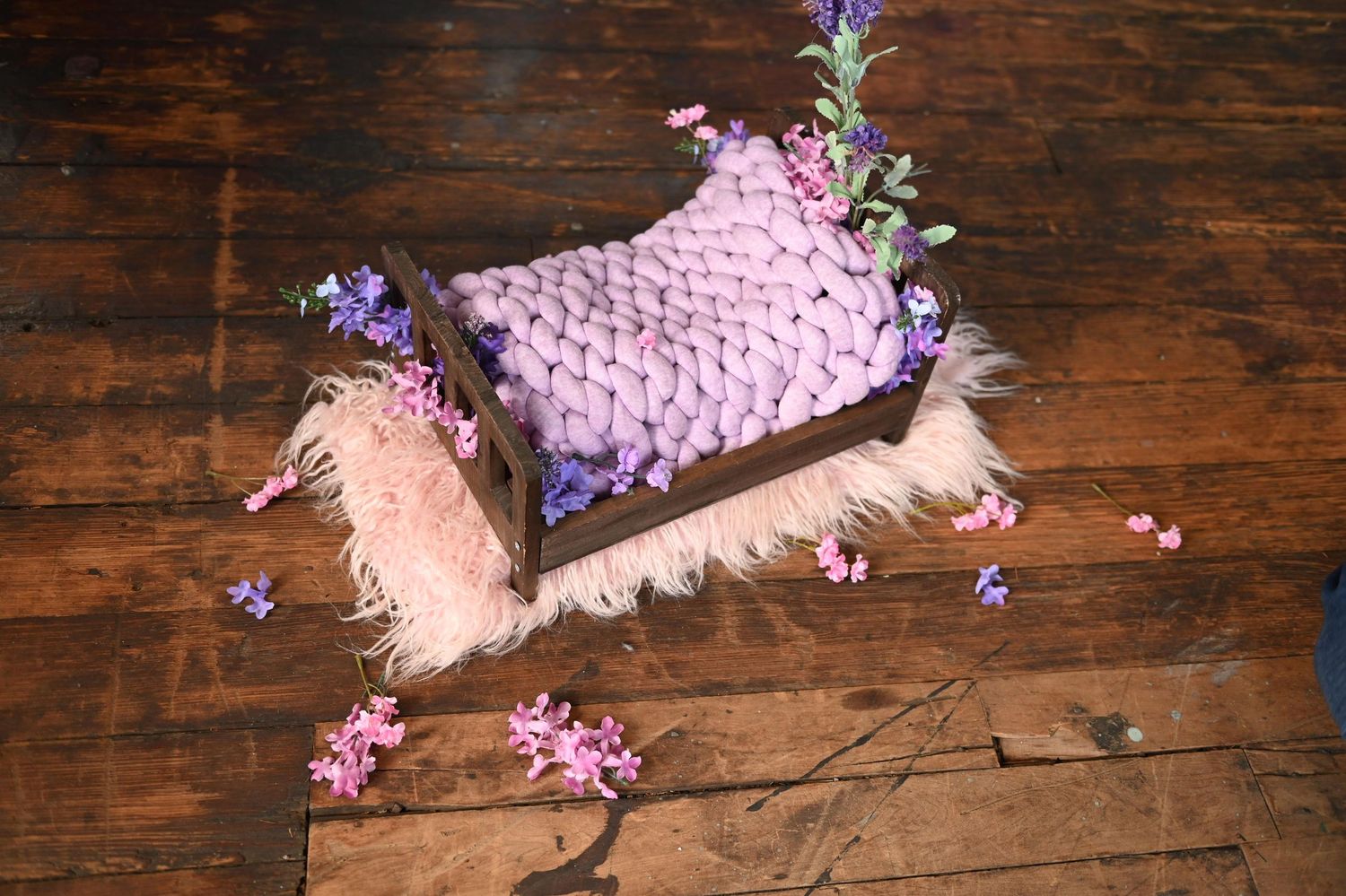 Small pink knitted pillow on wooden floor with scattered purple and pink flowers and fluffy pink fabric.