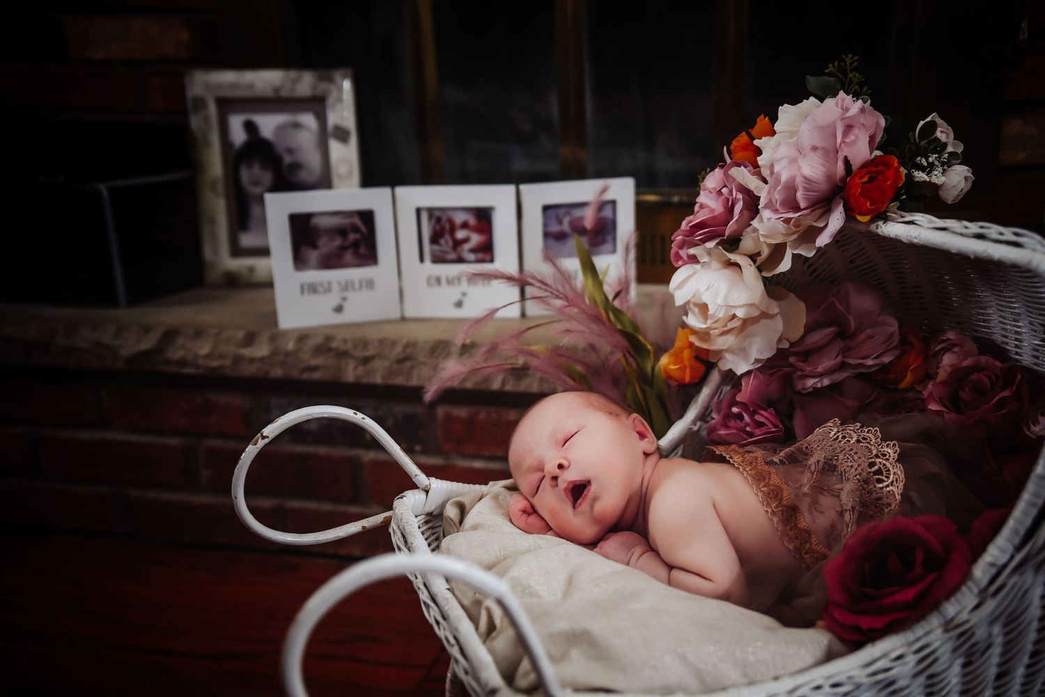 A newborn baby lies in a vintage white bassinet decorated with pink and burgundy flowers while photos are displayed behind.