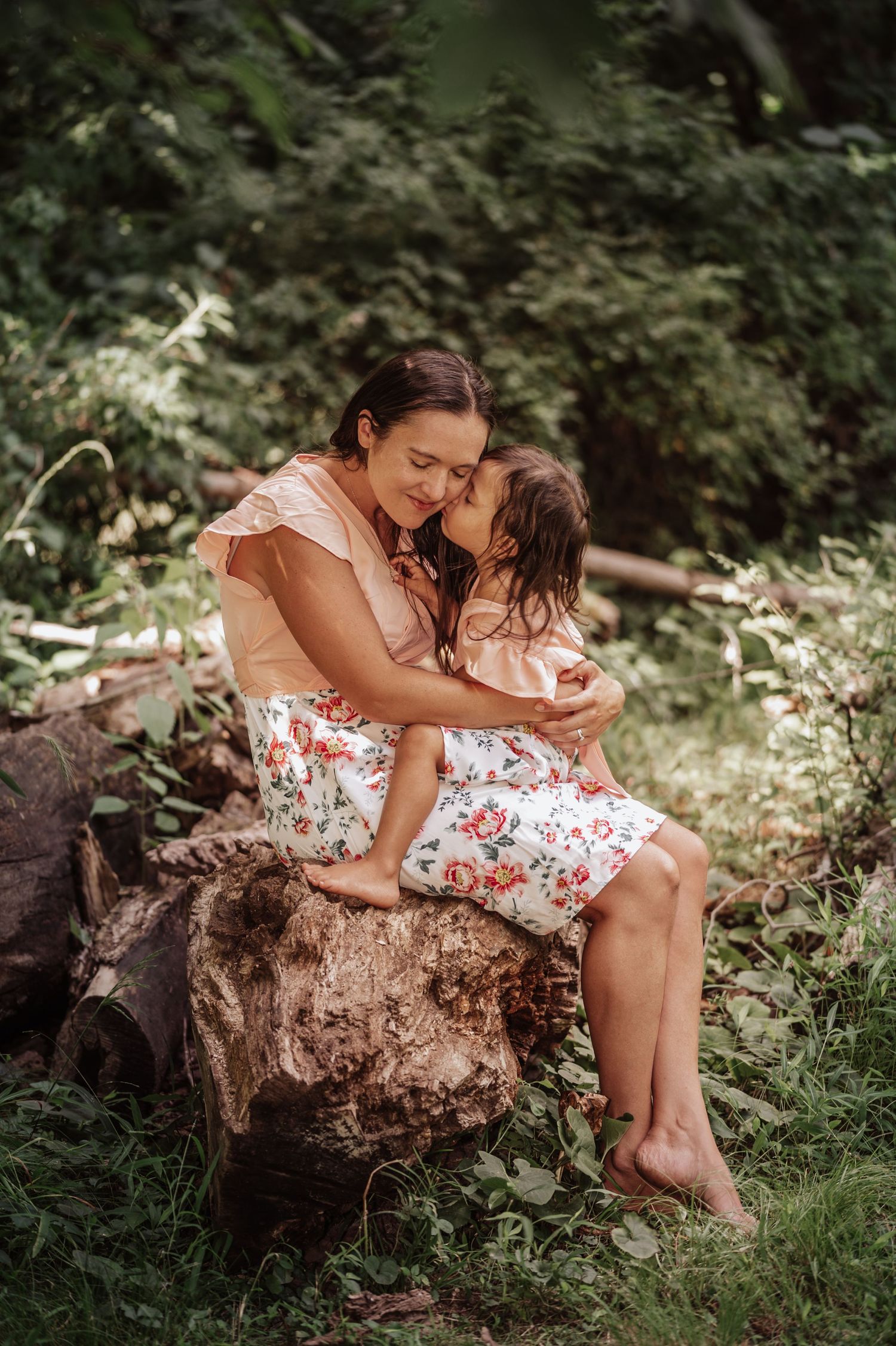 Two people in matching floral dresses share a tender moment while sitting on a large rock in a garden setting.