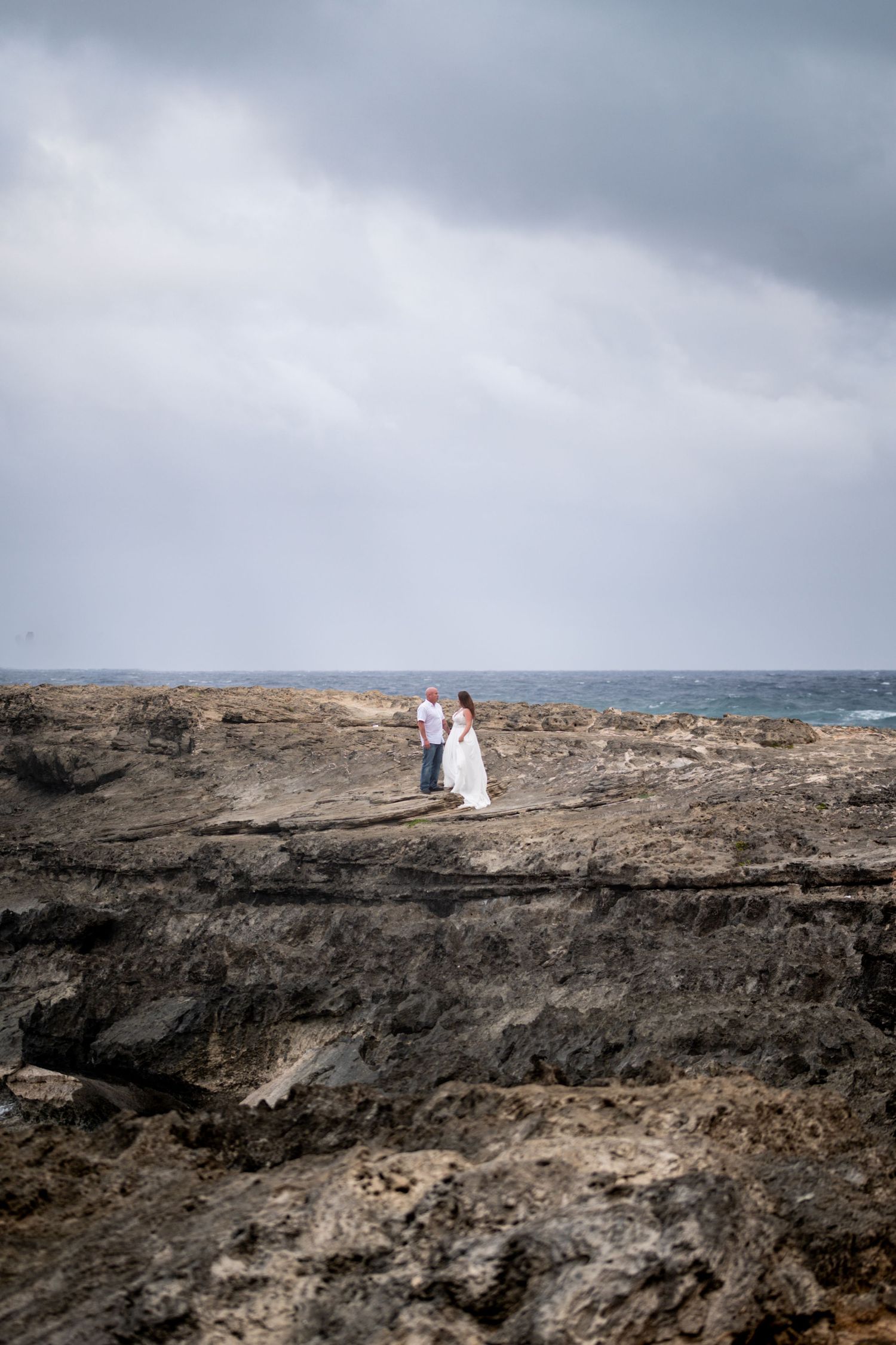 A couple stands on dramatic volcanic rock formations overlooking the ocean on a cloudy day.