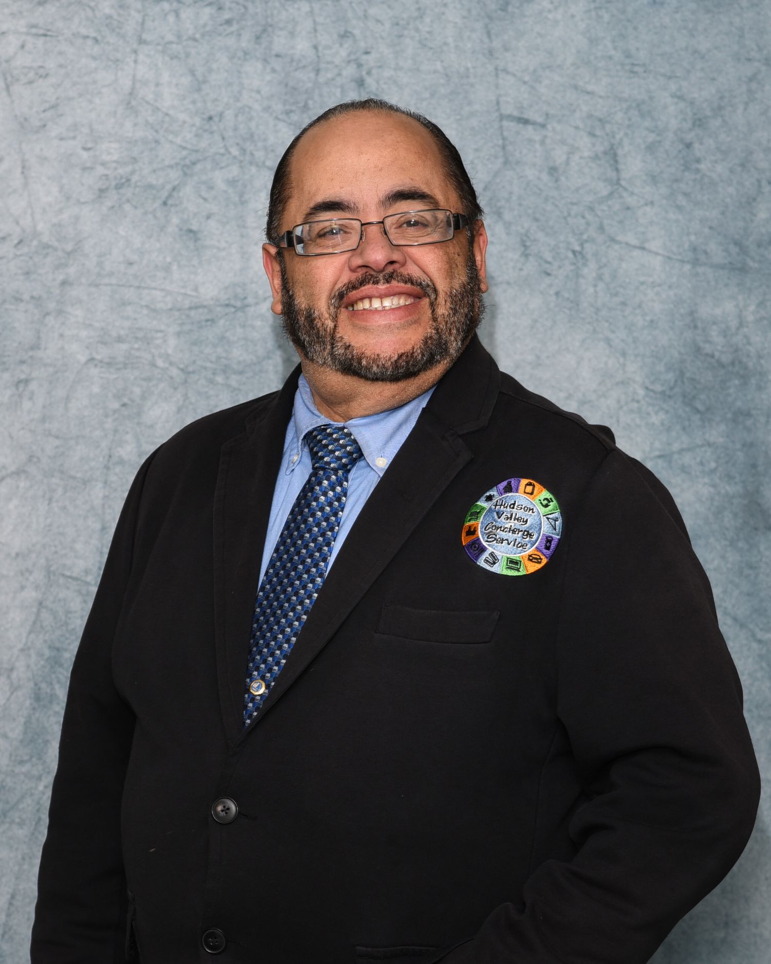 Professional headshot of someone in a dark suit and blue tie wearing glasses, with a colorful pin on their lapel.