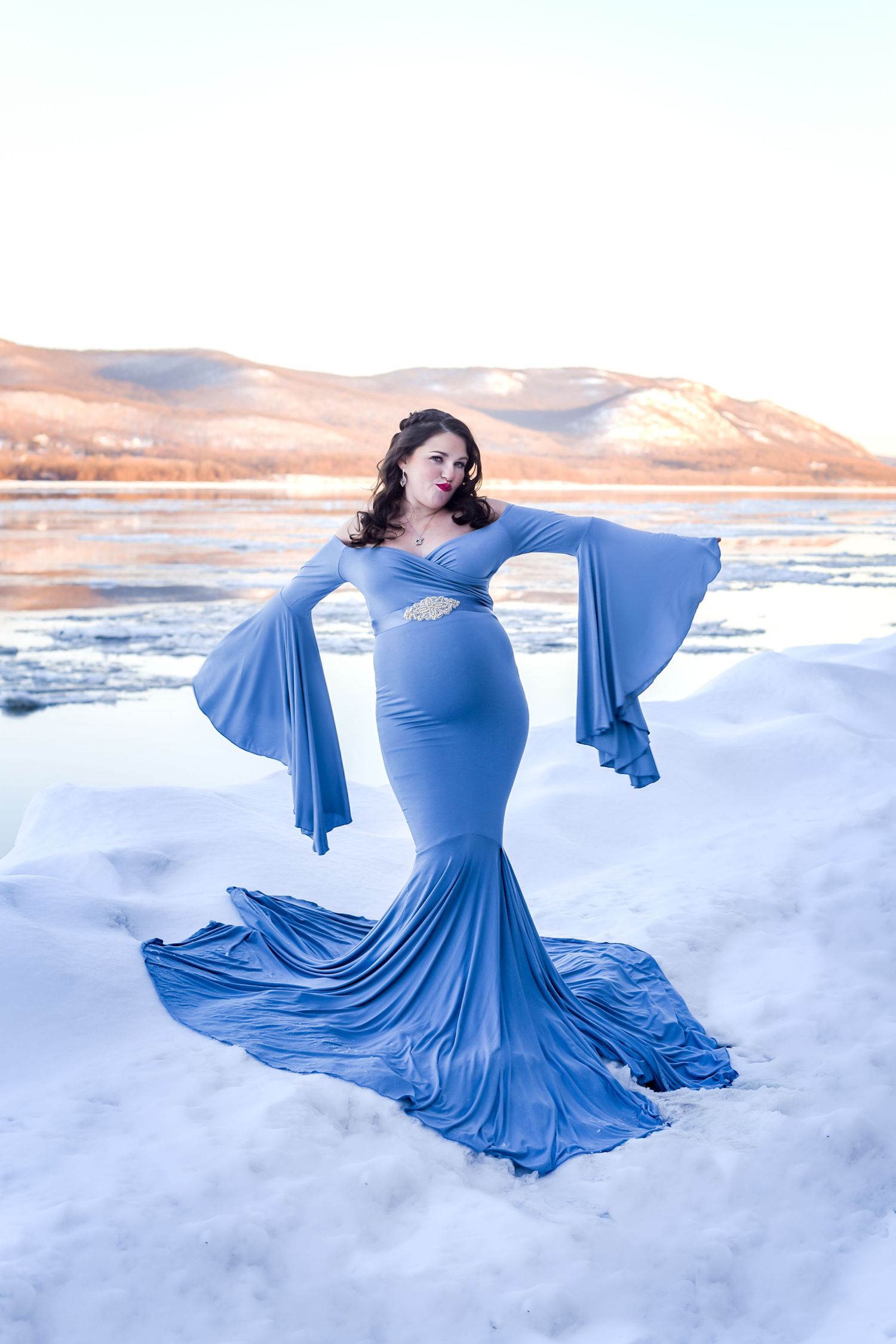 A pregnant woman in a flowing blue dress poses elegantly on a snowy landscape with mountains in the background.