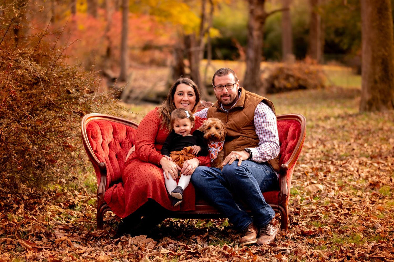 A red vintage couch sits outdoors in autumn leaves with people and dogs enjoying a fall family photoshoot.