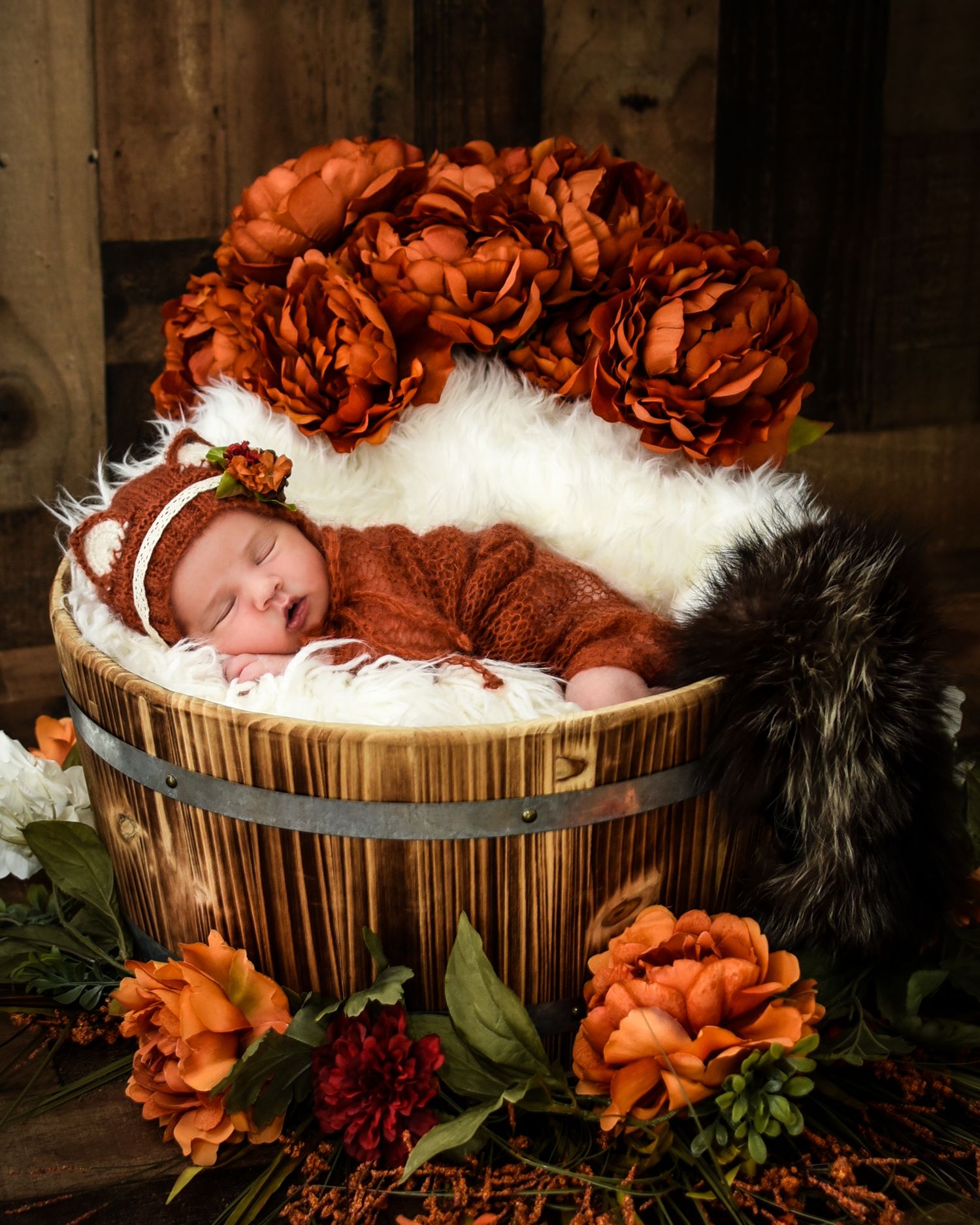 A sleeping baby in an orange outfit nestles in a rustic wooden basket surrounded by autumn flowers and white fur.