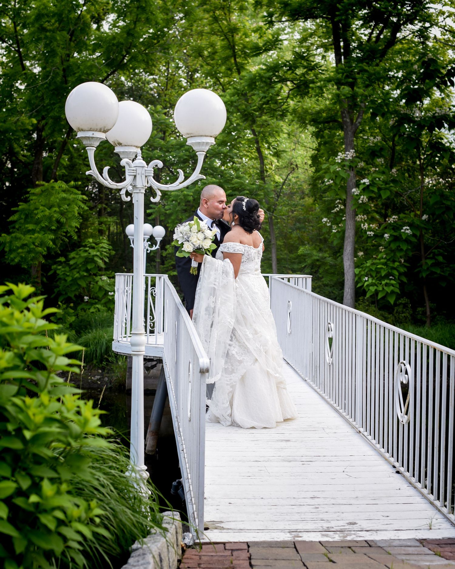Newlyweds share romantic moment on white bridge with ornate lamp post in garden setting.