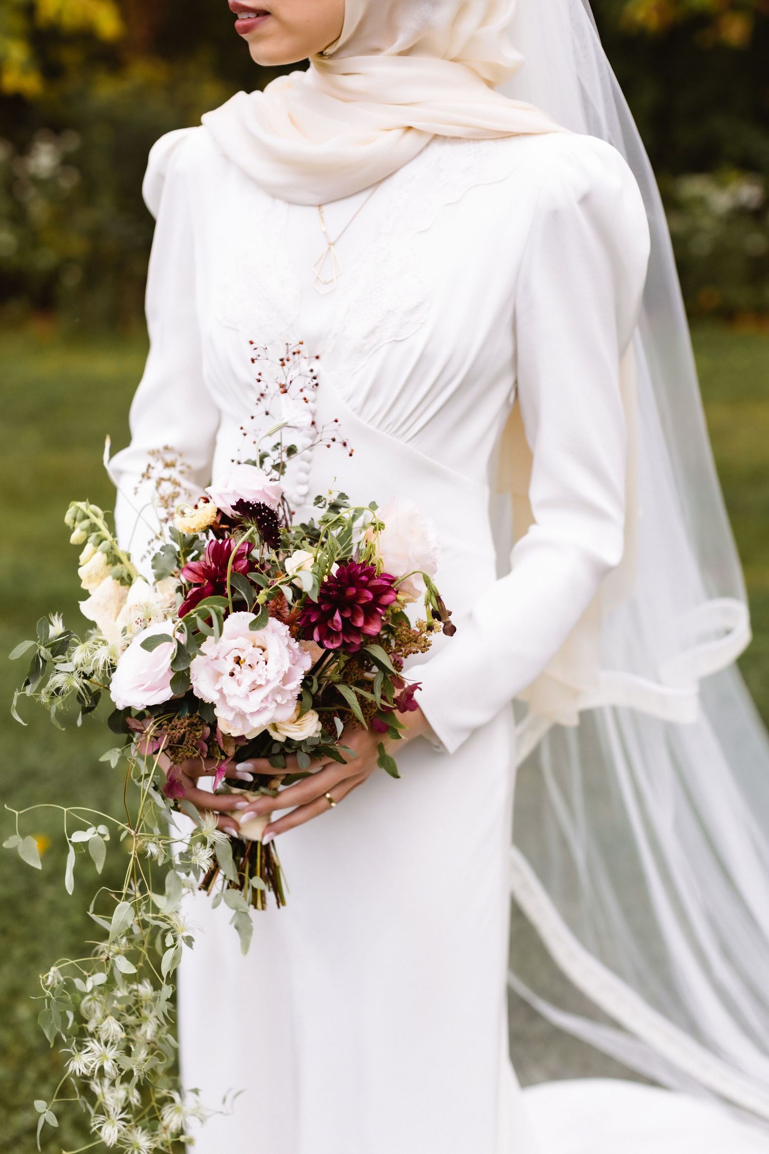 Elegant long-sleeved white wedding dress with veil and romantic burgundy floral bouquet featuring peonies and greenery.