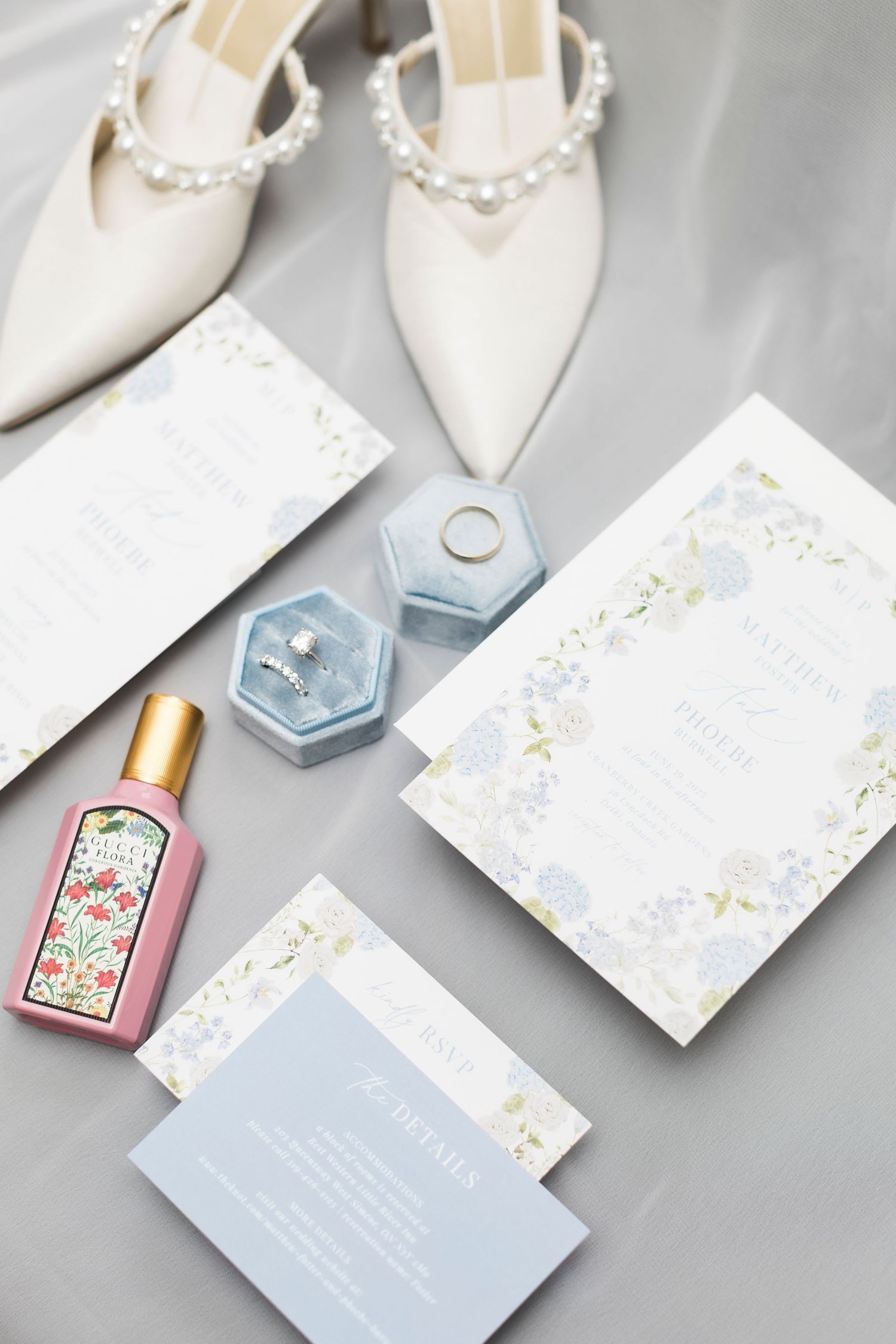 Elegant wedding details flatlay with white heels, floral stationery, perfume bottle and ring box in soft blue tones.