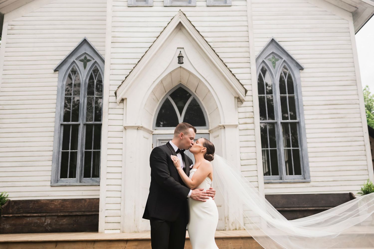 A couple shares a romantic moment in front of a white wooden church with gothic windows and a pointed archway entrance.