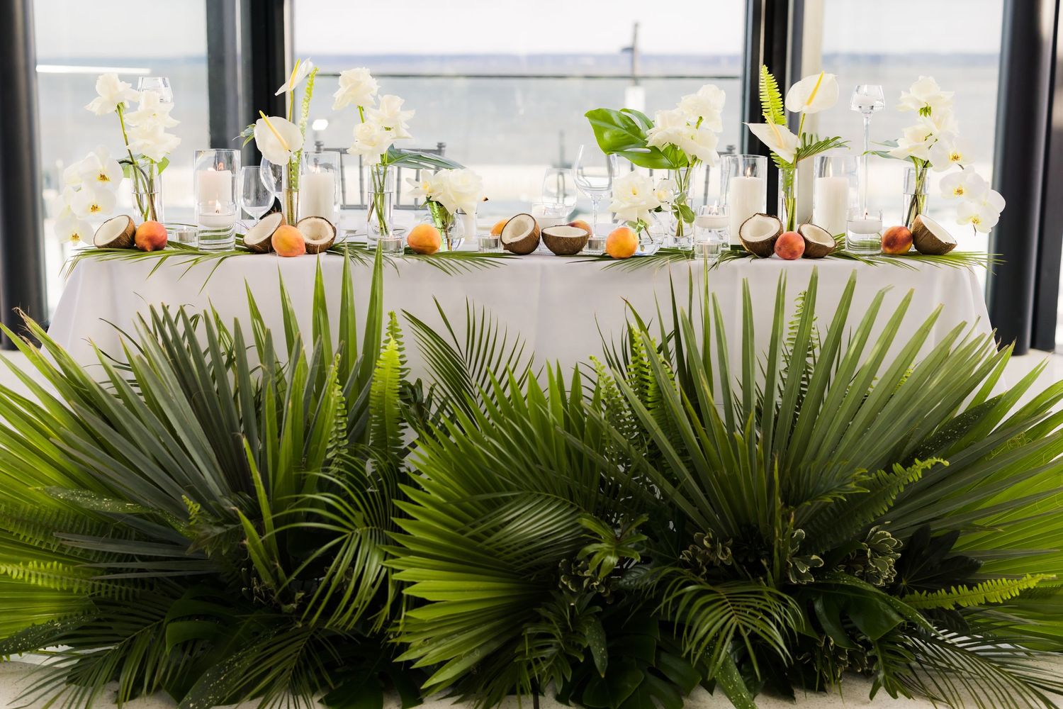 Modern wedding reception table with tropical palm decor and white floral centerpieces.