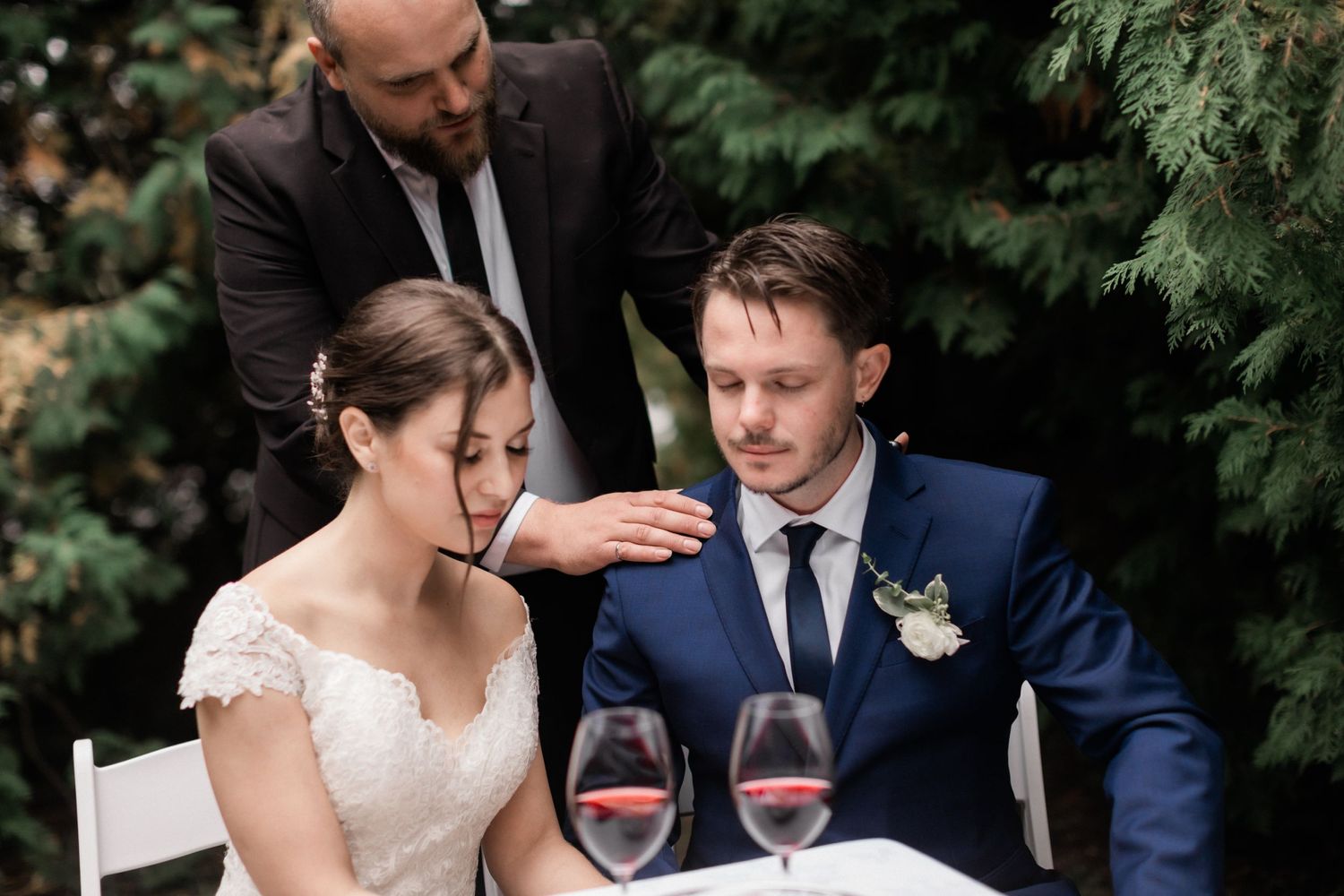 Newlyweds sit at an outdoor table with wine glasses while an officiant stands behind them during an intimate ceremony.