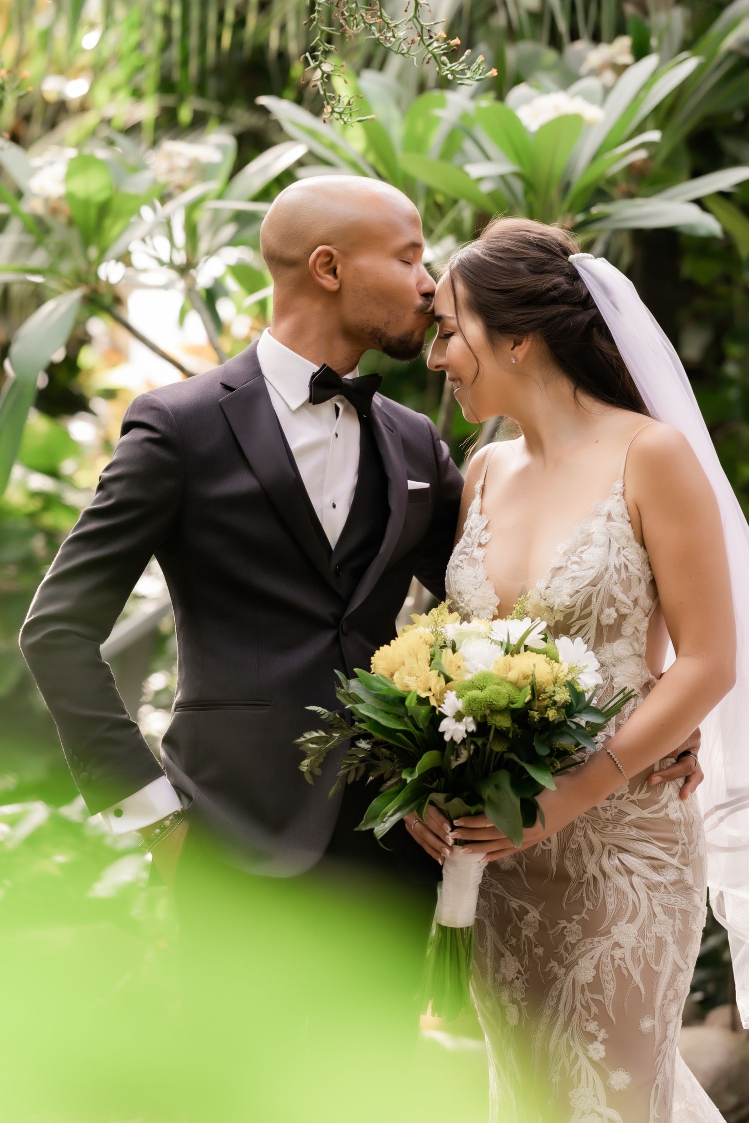 Romantic wedding portrait of couple with bride holding yellow bouquet in tropical garden setting.