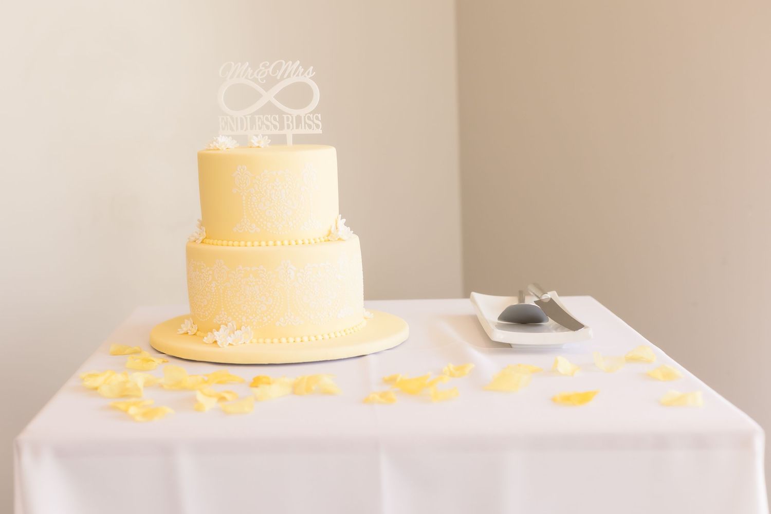 Two-tier pale yellow wedding cake with delicate lace details and monogram topper on white tablecloth.