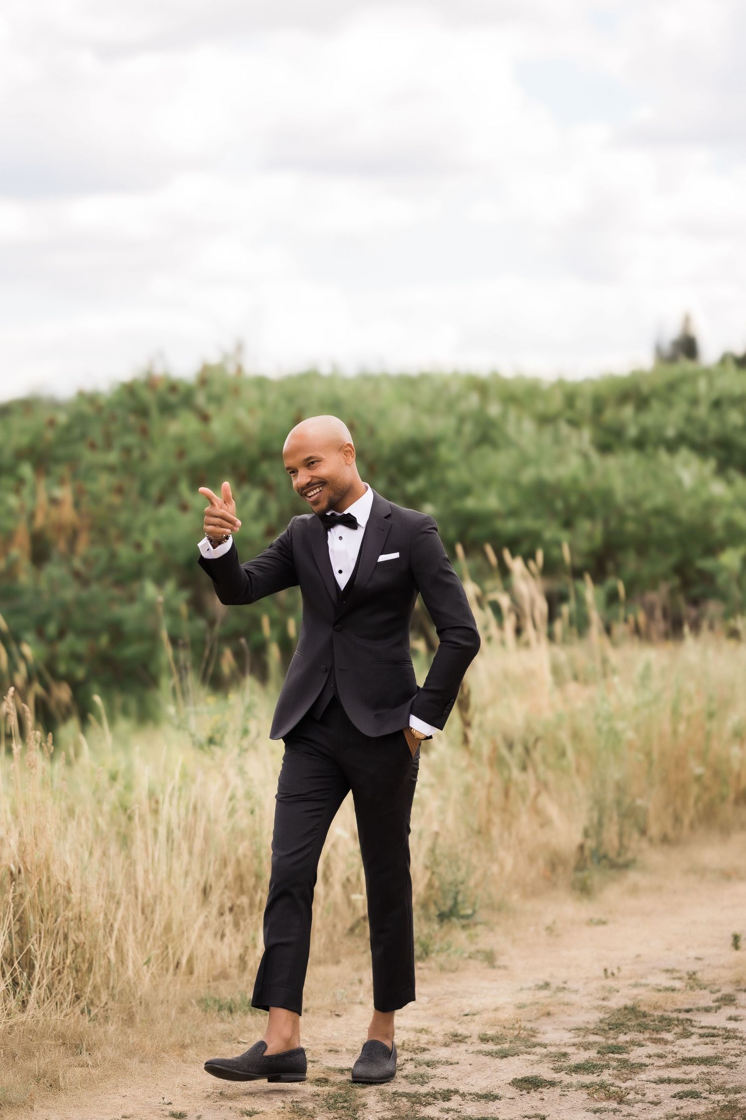 Well-dressed person in black tuxedo poses confidently on dirt path surrounded by tall grass.