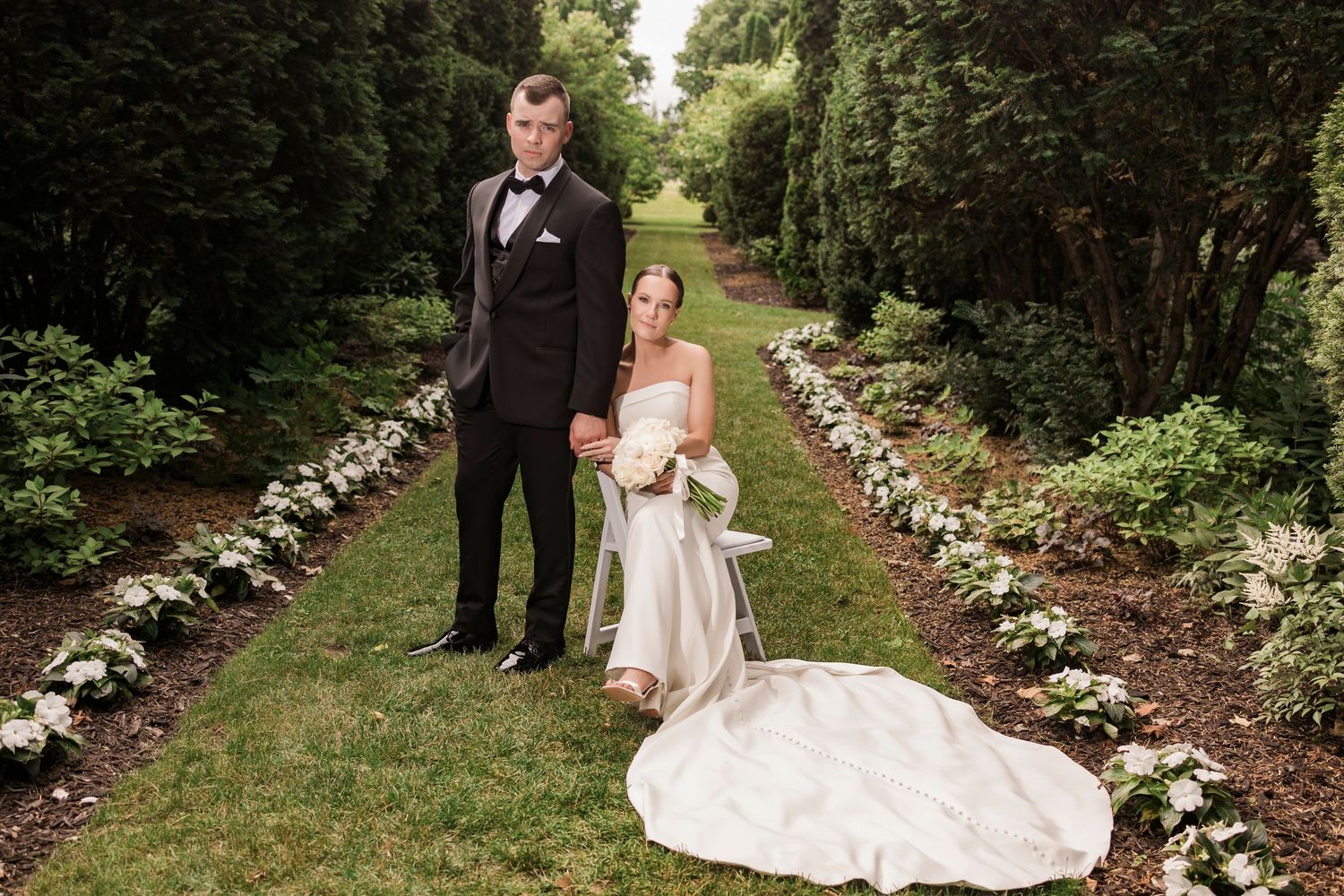 Wedding couple poses in garden path lined with white flowers.
