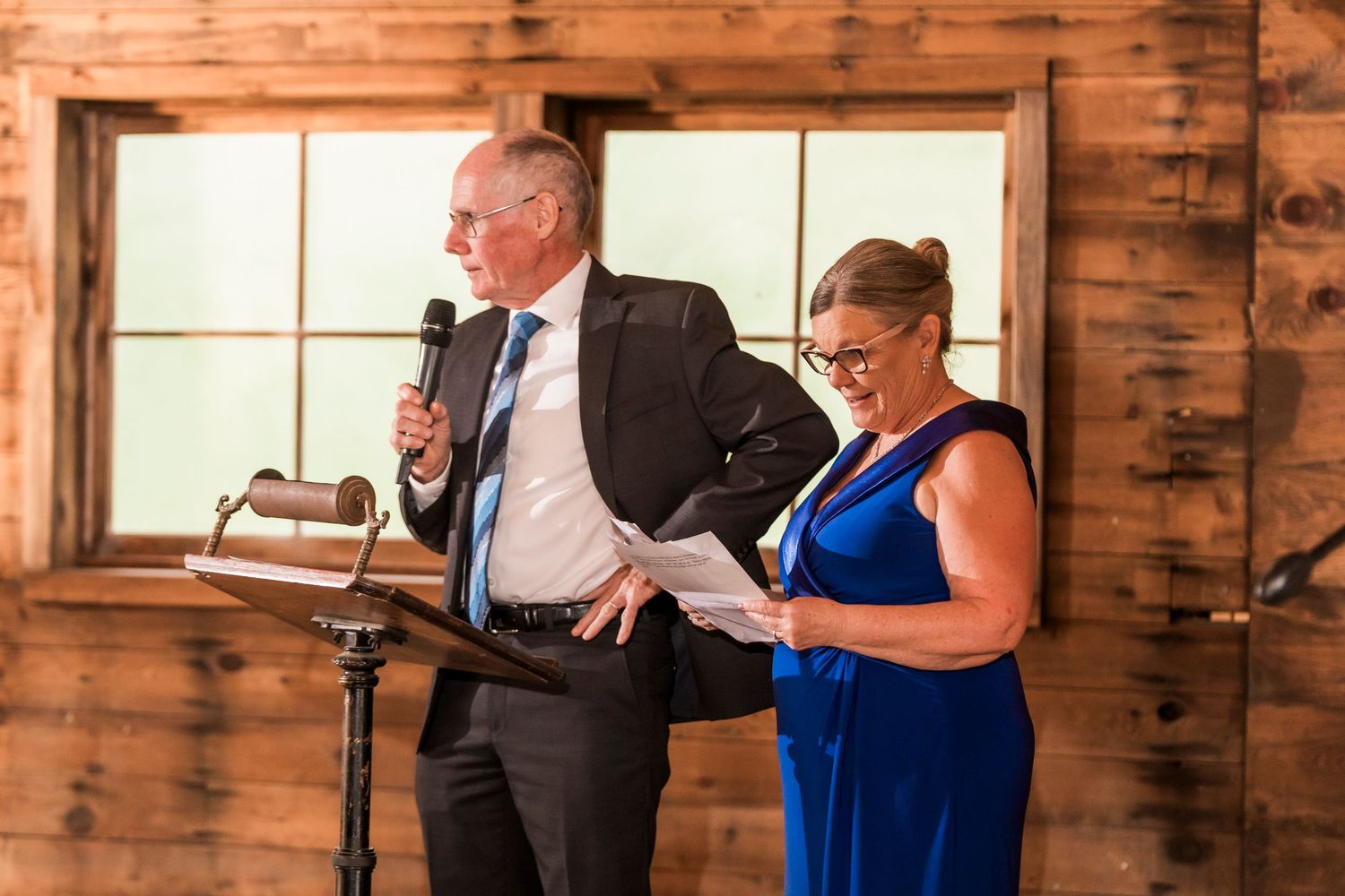Wedding guests gather at rustic tables for dinner in a warm wooden barn setting with bright windows.