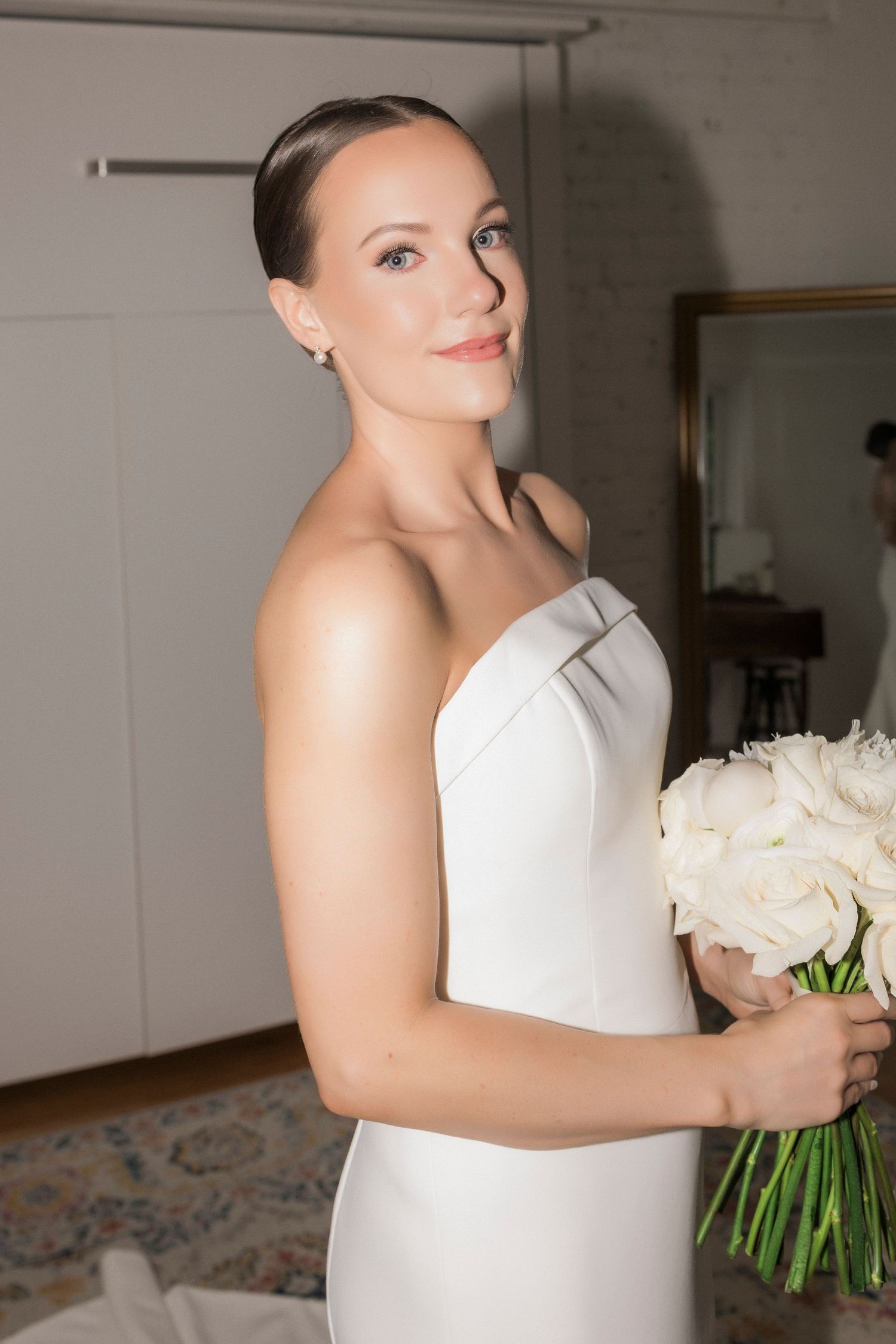 Close-up portrait of bride in simple white gown holding white rose bouquet against neutral background.