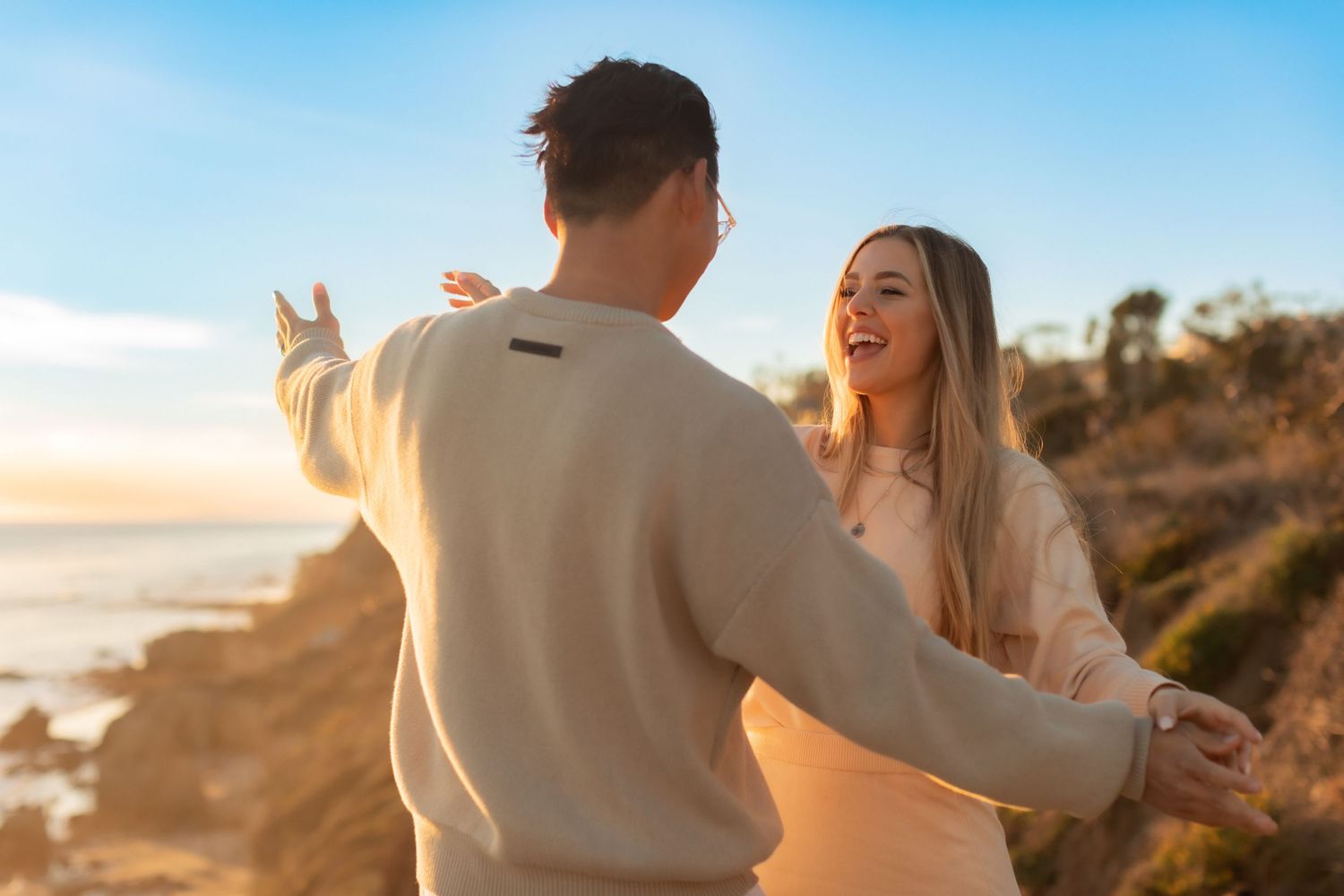 Couple enjoying playful moment on coastal cliff edge during sunset.