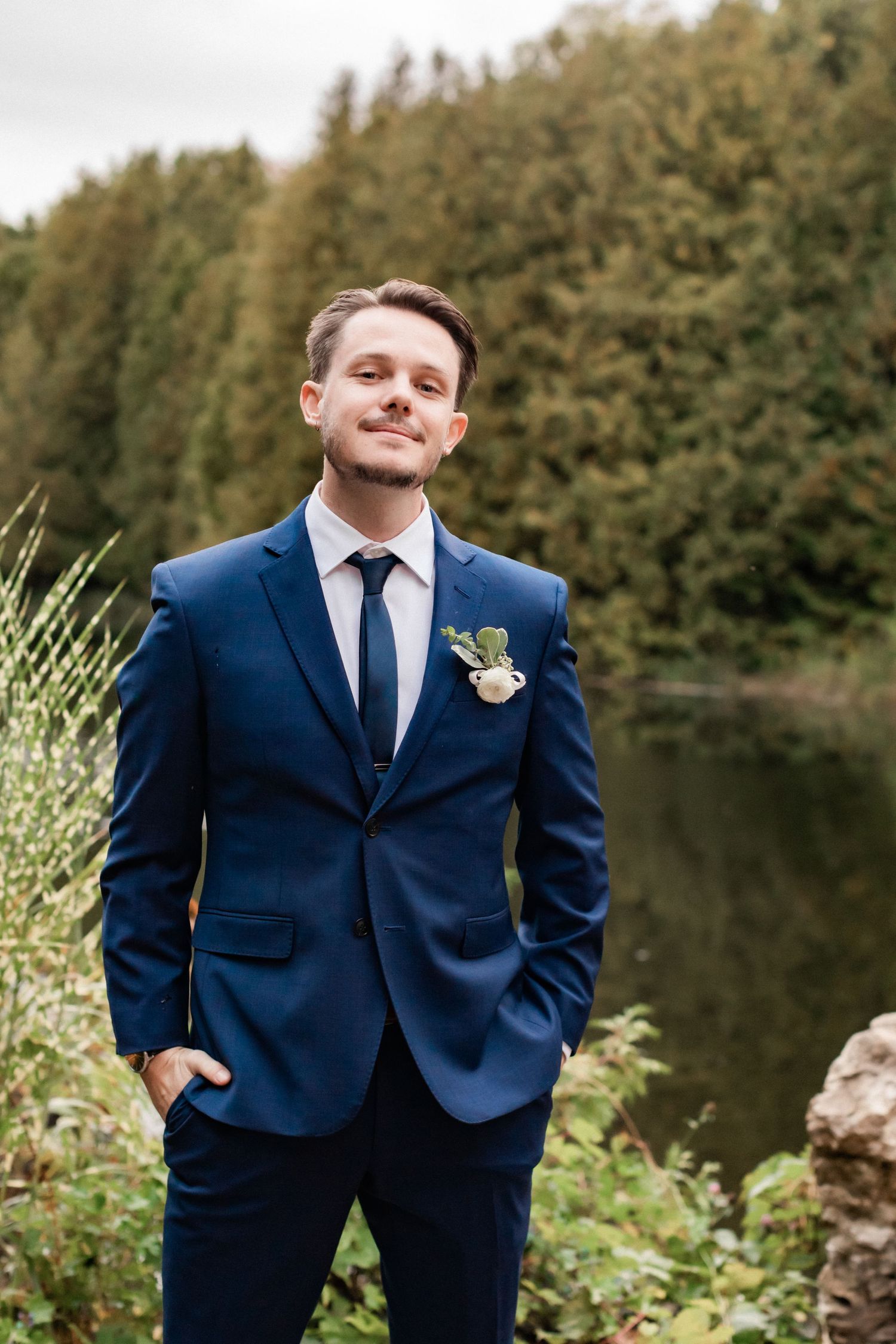 A groom in a navy blue suit poses confidently by a lake with forest backdrop.