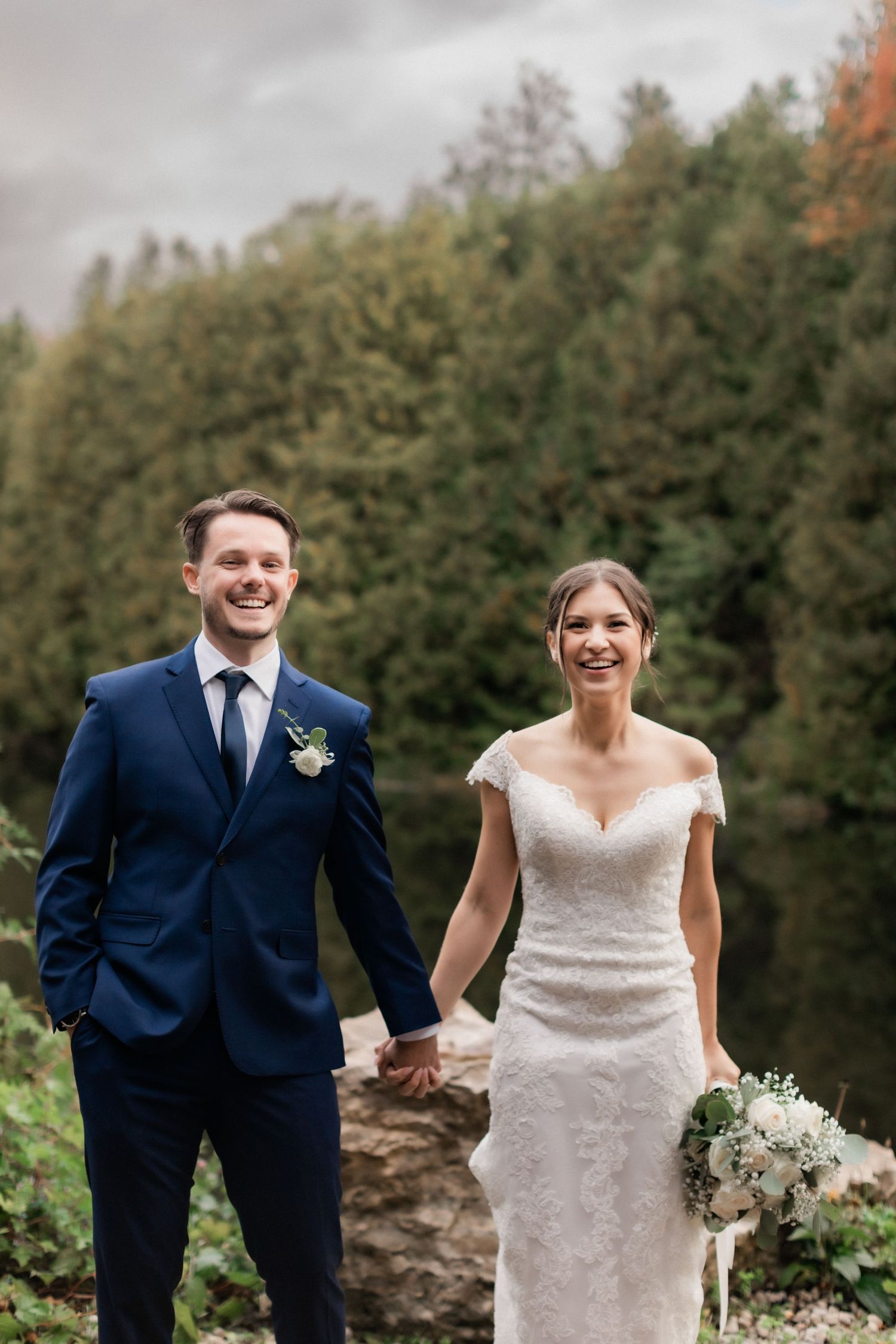 Newlyweds smile while standing together in front of an autumn forest landscape.